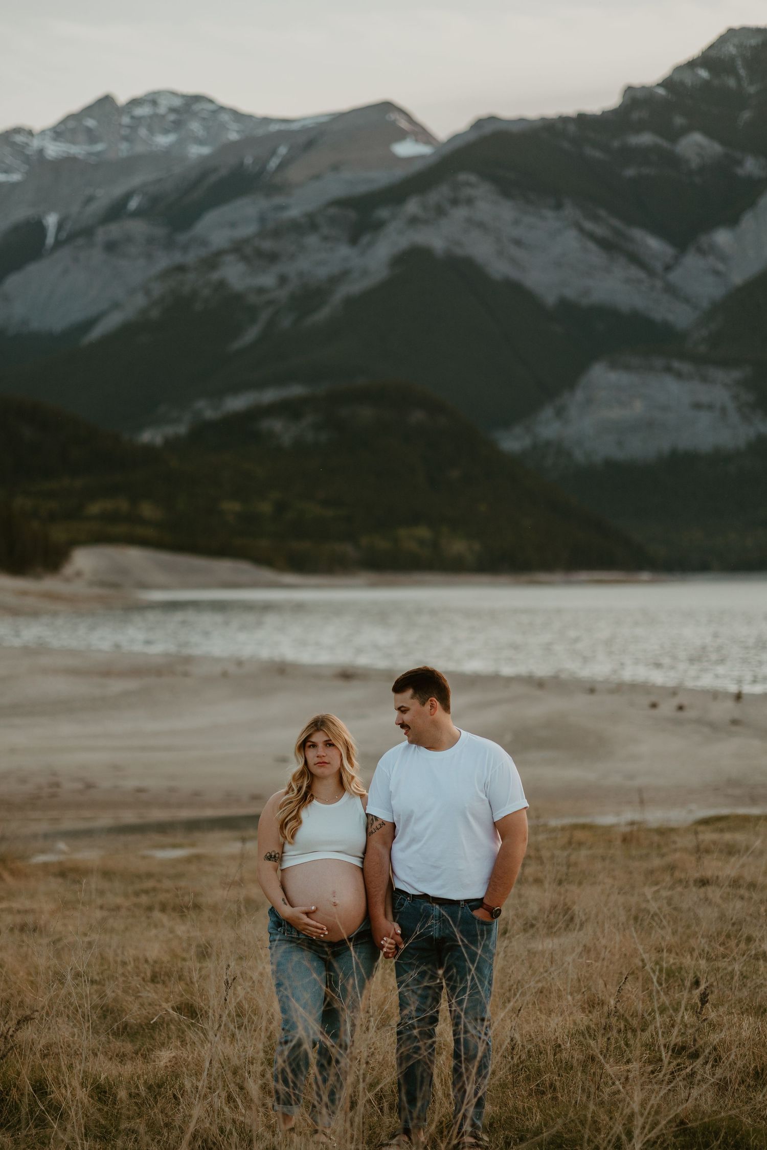 Couple stands together in field with dramatic mountain landscape behind them.