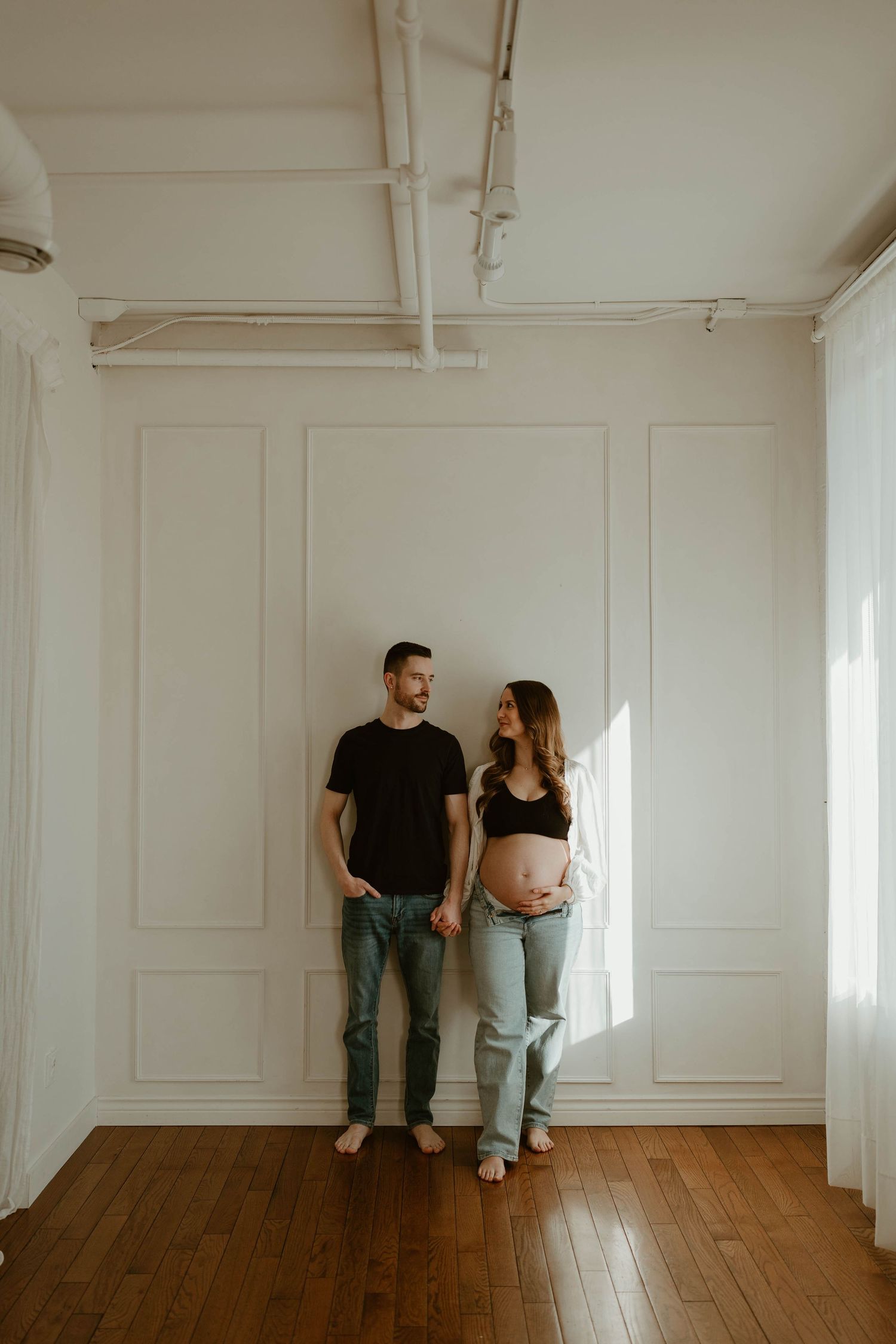 A couple stands against a white wall, the woman visibly pregnant, both wearing jeans in a bright minimalist room.