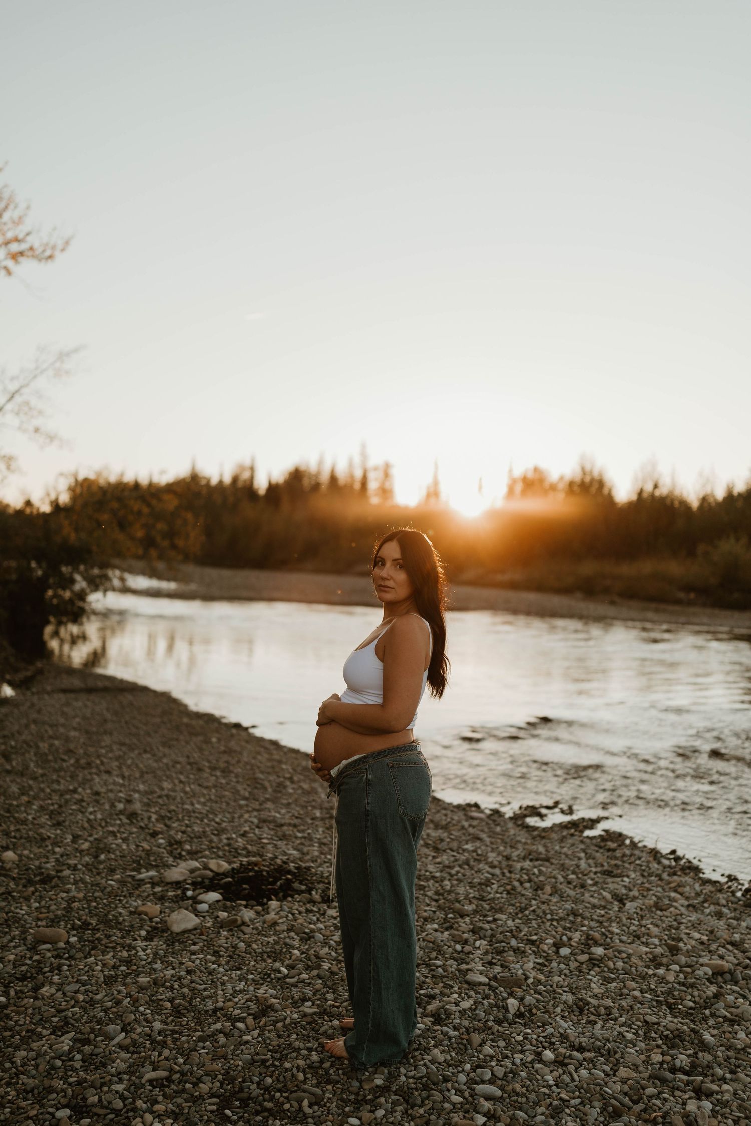 Pregnant woman in white crop top and teal pants cradling baby bump by a river at golden sunset.