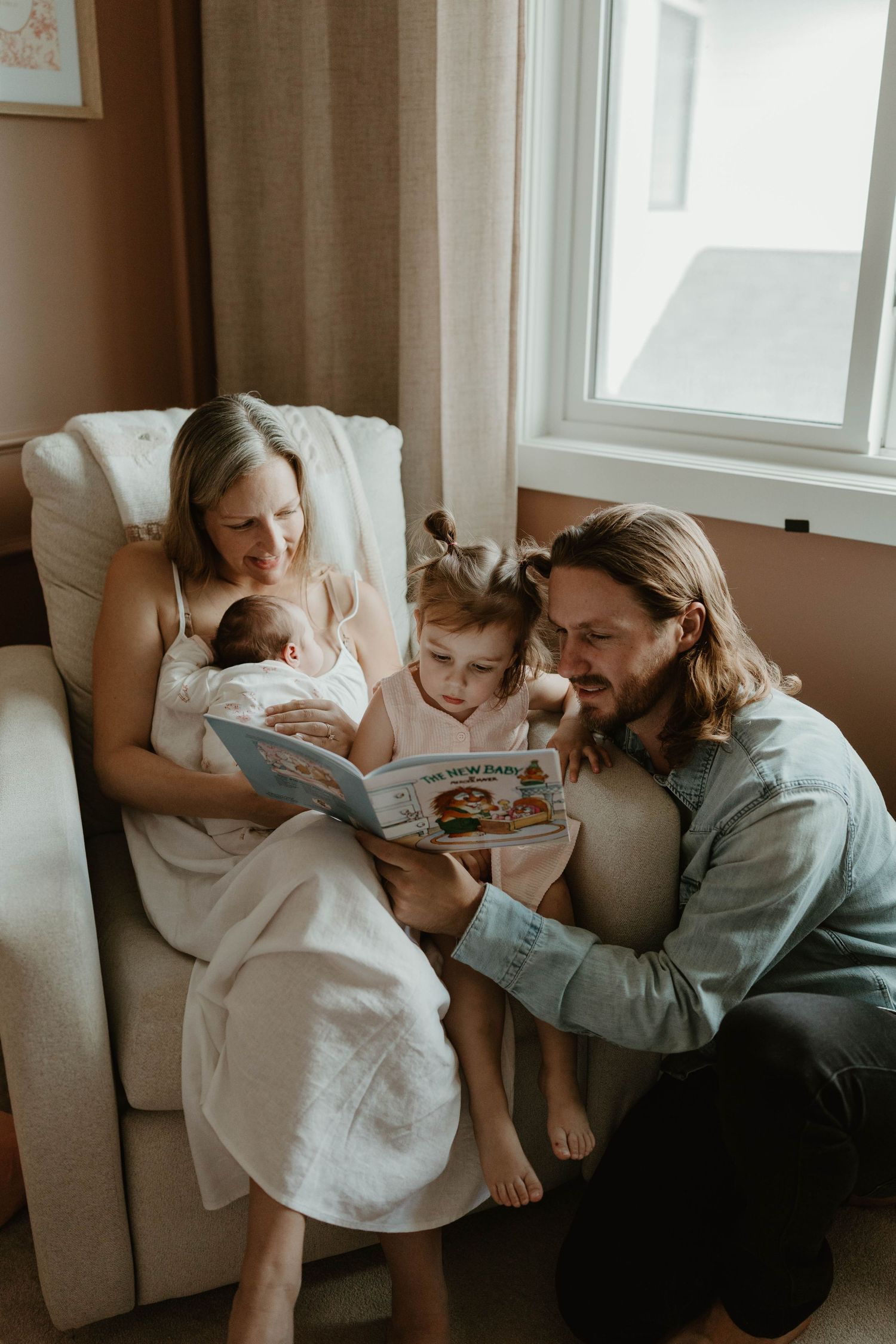 Family of four reads a picture book together in a cozy armchair near a window, with a newborn baby and toddler.