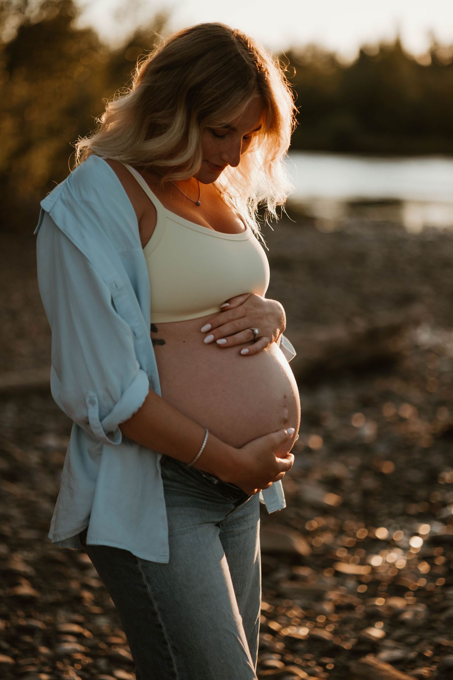 Pregnant woman cradling her baby bump at golden hour on a rocky beach, wearing a white crop top and open blue shirt.