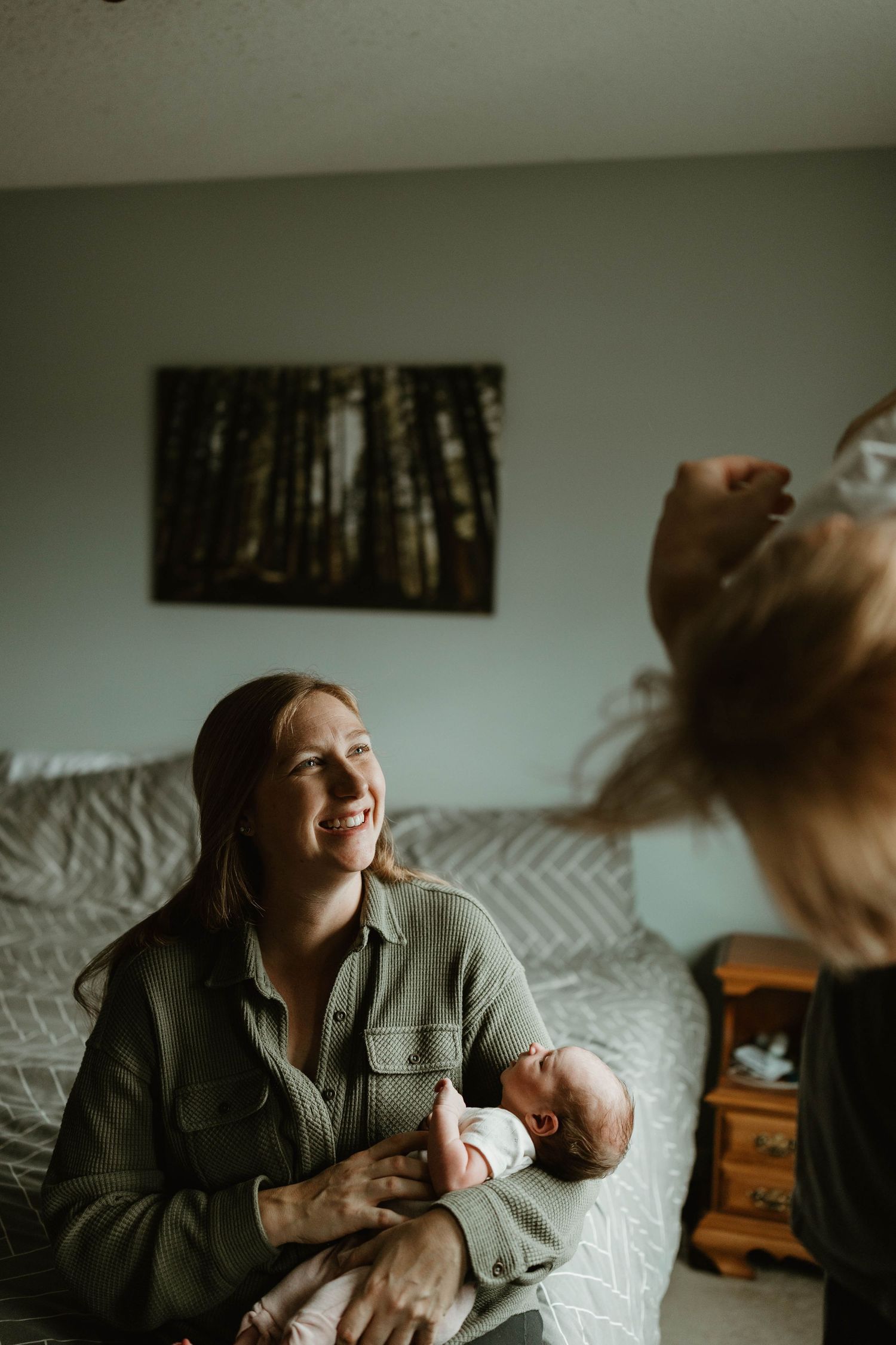 A smiling mother holds her newborn baby while an older child plays nearby in a cozy bedroom.