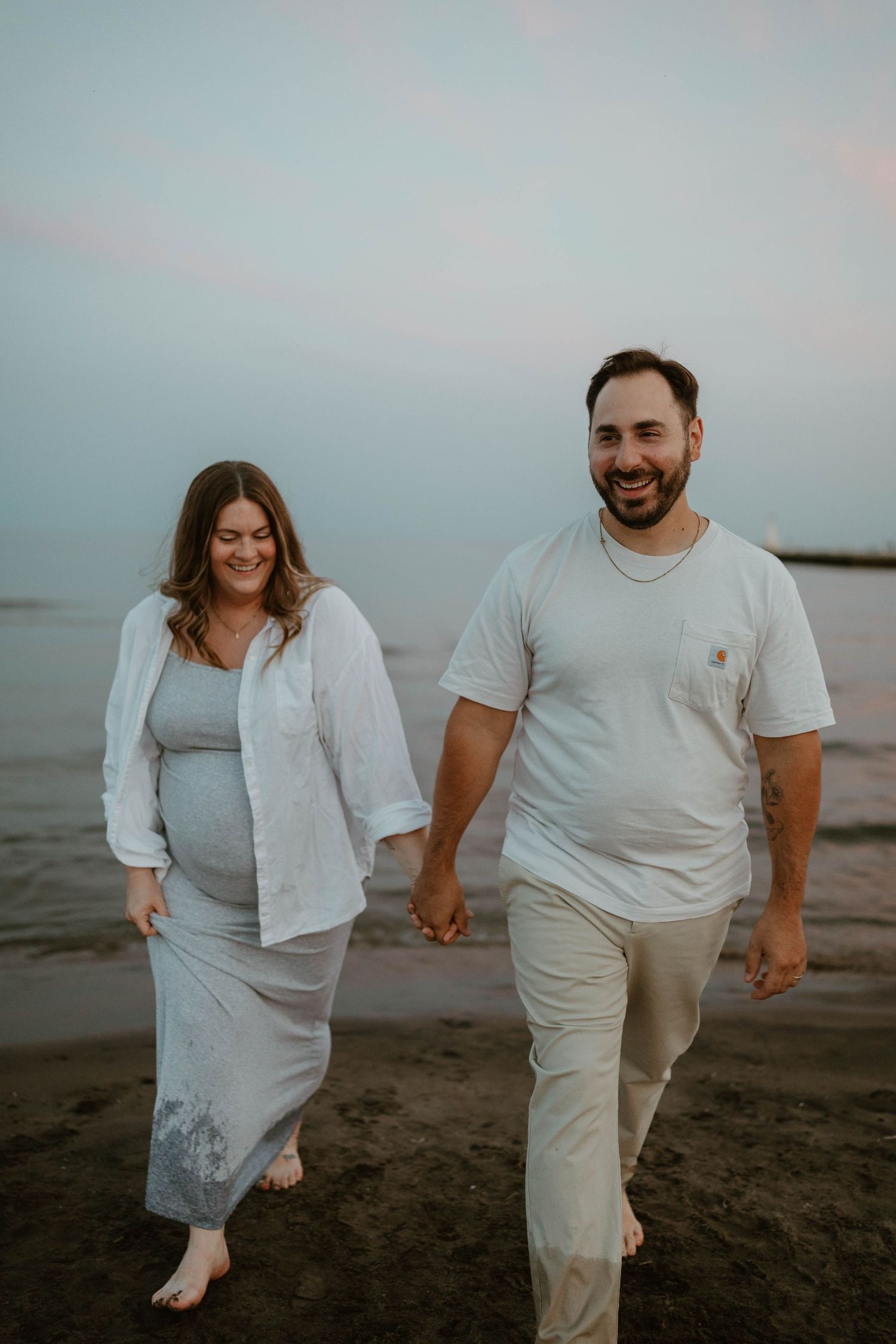 Pregnant couple holding hands and walking barefoot on a beach at sunset, smiling joyfully.