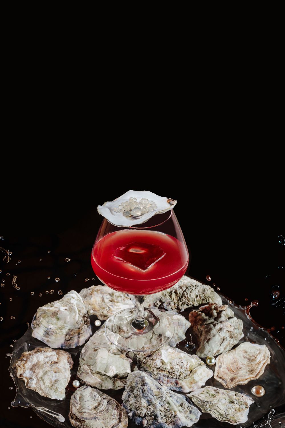 Red cocktail in stemmed glass perched atop pile of fresh oysters against dark background.