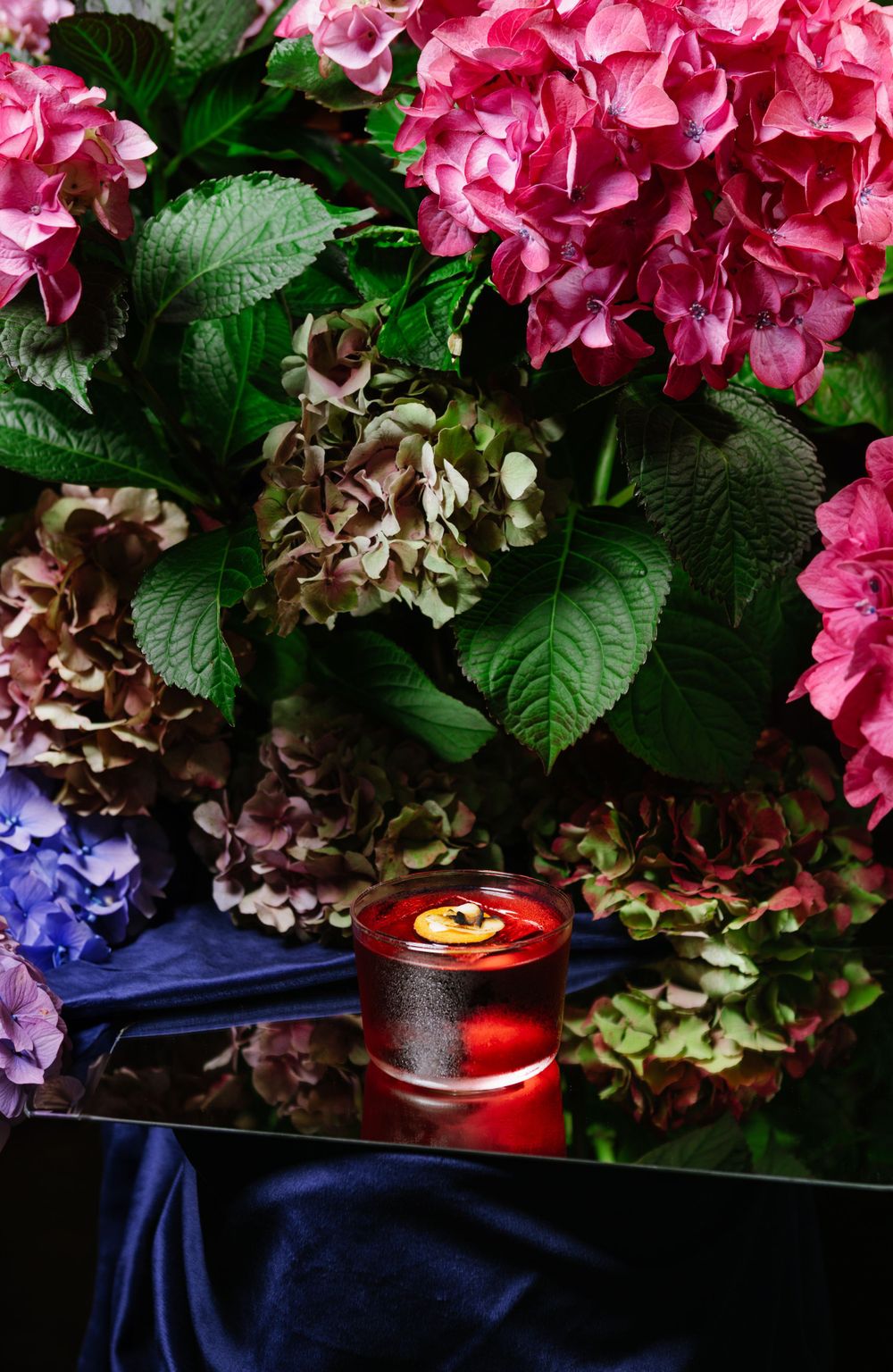 Pink and purple hydrangeas with green leaves arranged around glowing red candle.