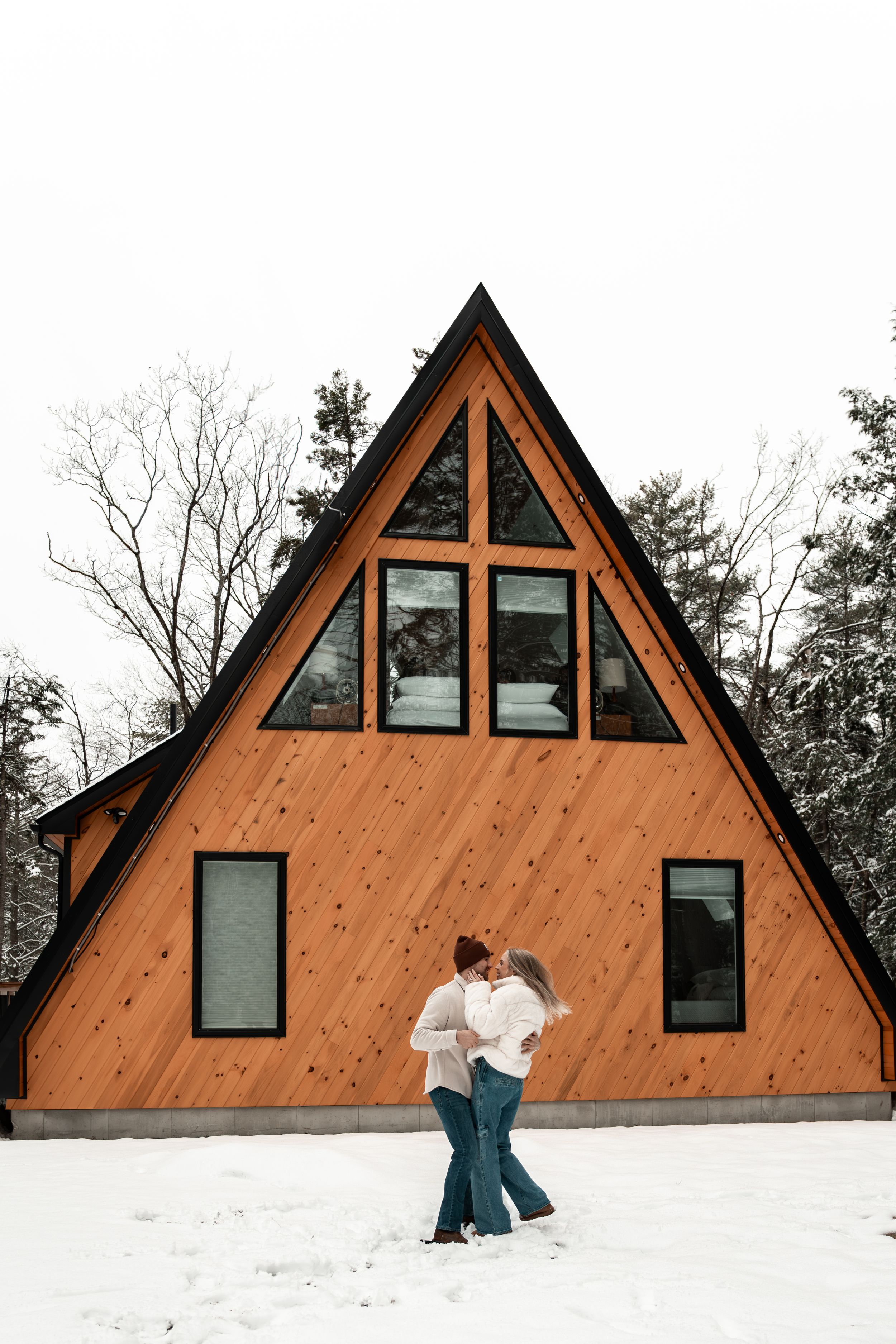 A-frame wooden cabin with large triangular windows stands in snowy landscape while couple dances in front in a winter wonderland