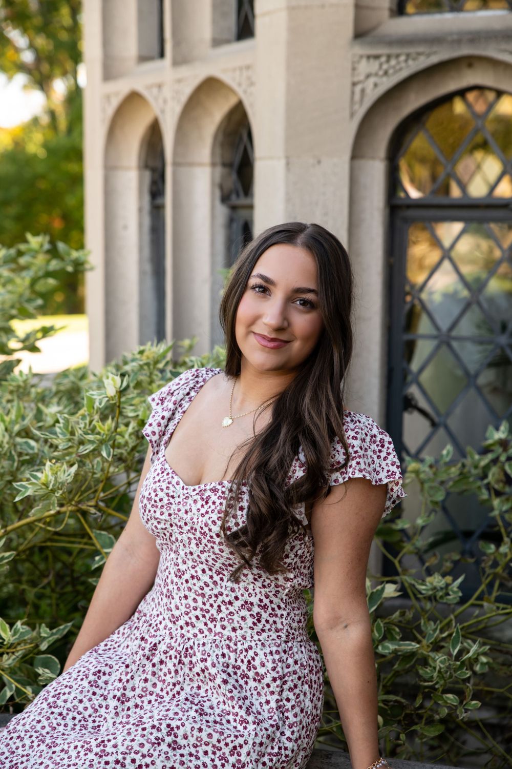 Brunette young woman in floral dress sitting gracefully in front of a stone gothic-style building.