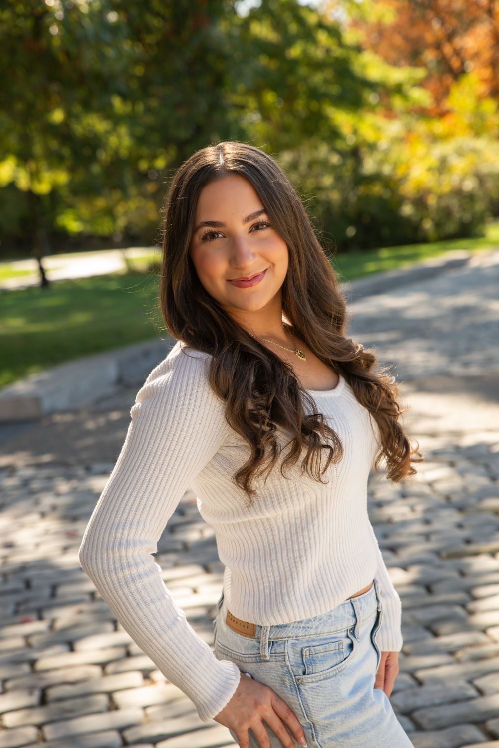 Smiling young woman in white ribbed top and jeans posing on cobblestone path in autumn park.