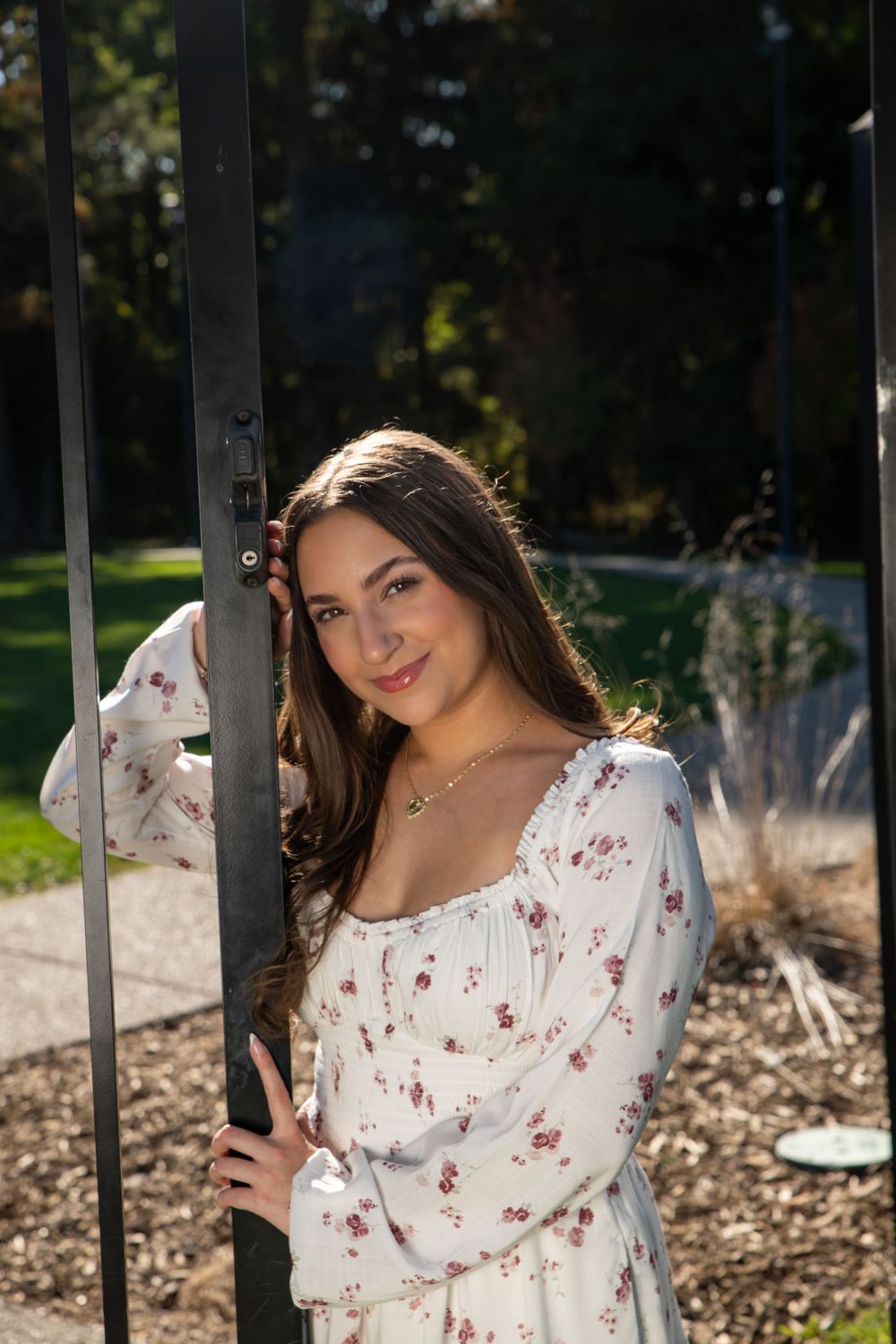 Young woman in floral dress leaning against black iron gate in sunny park.
