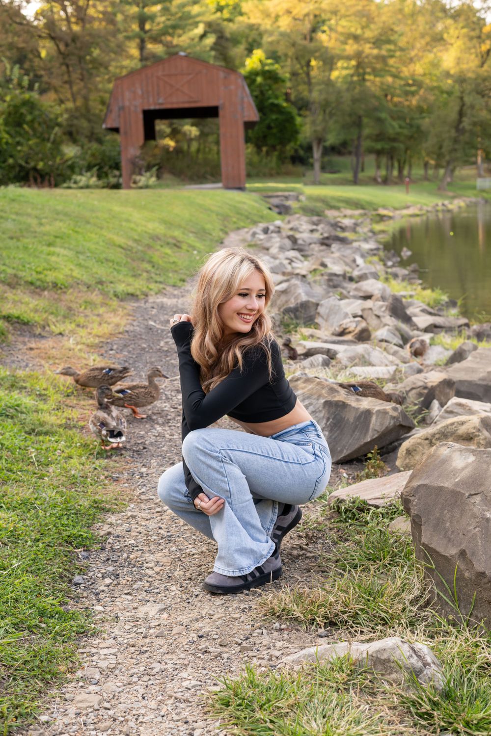 Smiling blonde girl crouching near ducks by a rocky pond with covered bridge.