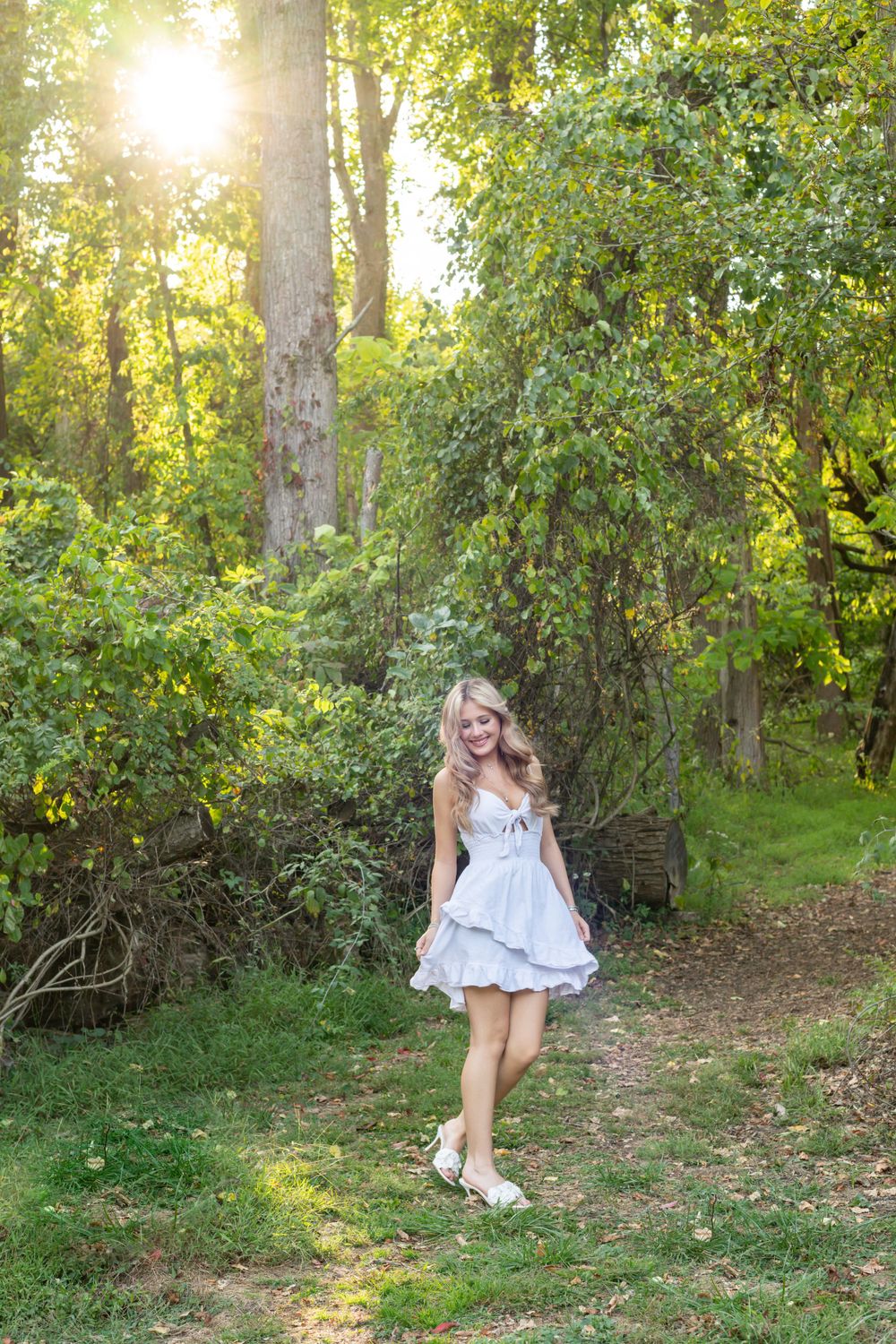 Blonde girl twirling in short white dress among lush green trees at sunset.