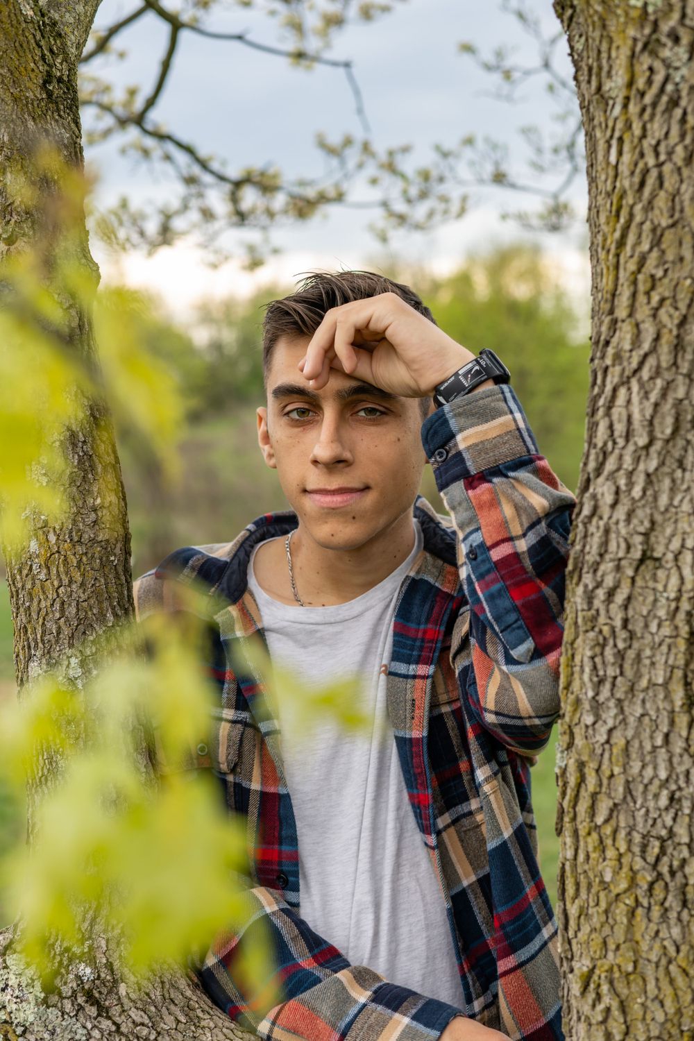 Young man in plaid shirt posing between two trees in a green park setting.