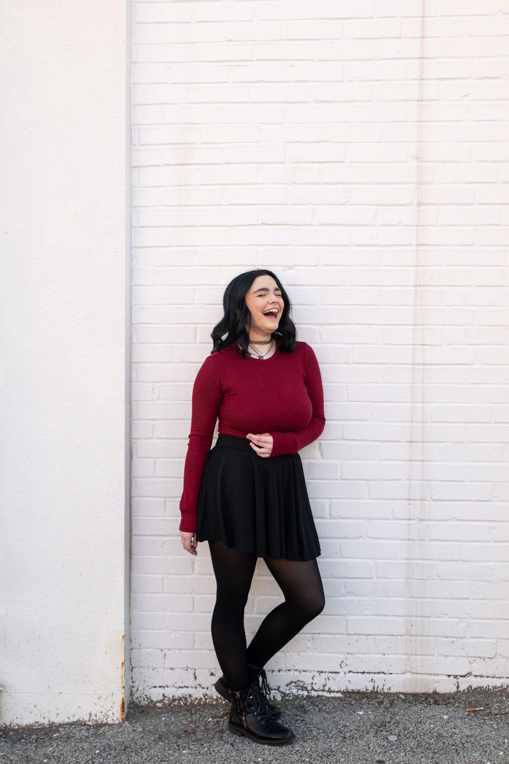 Young woman in red sweater and black skirt laughing against white brick wall.