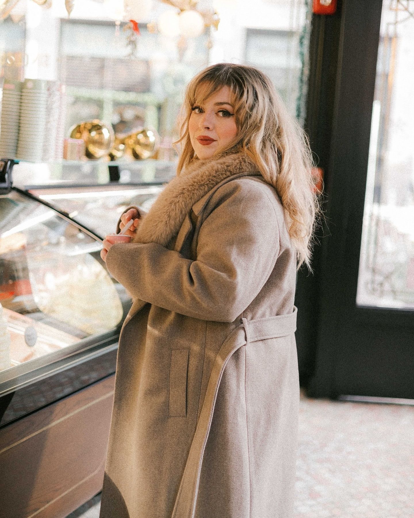 Person in a beige winter coat shopping at a store display window with holiday decorations.