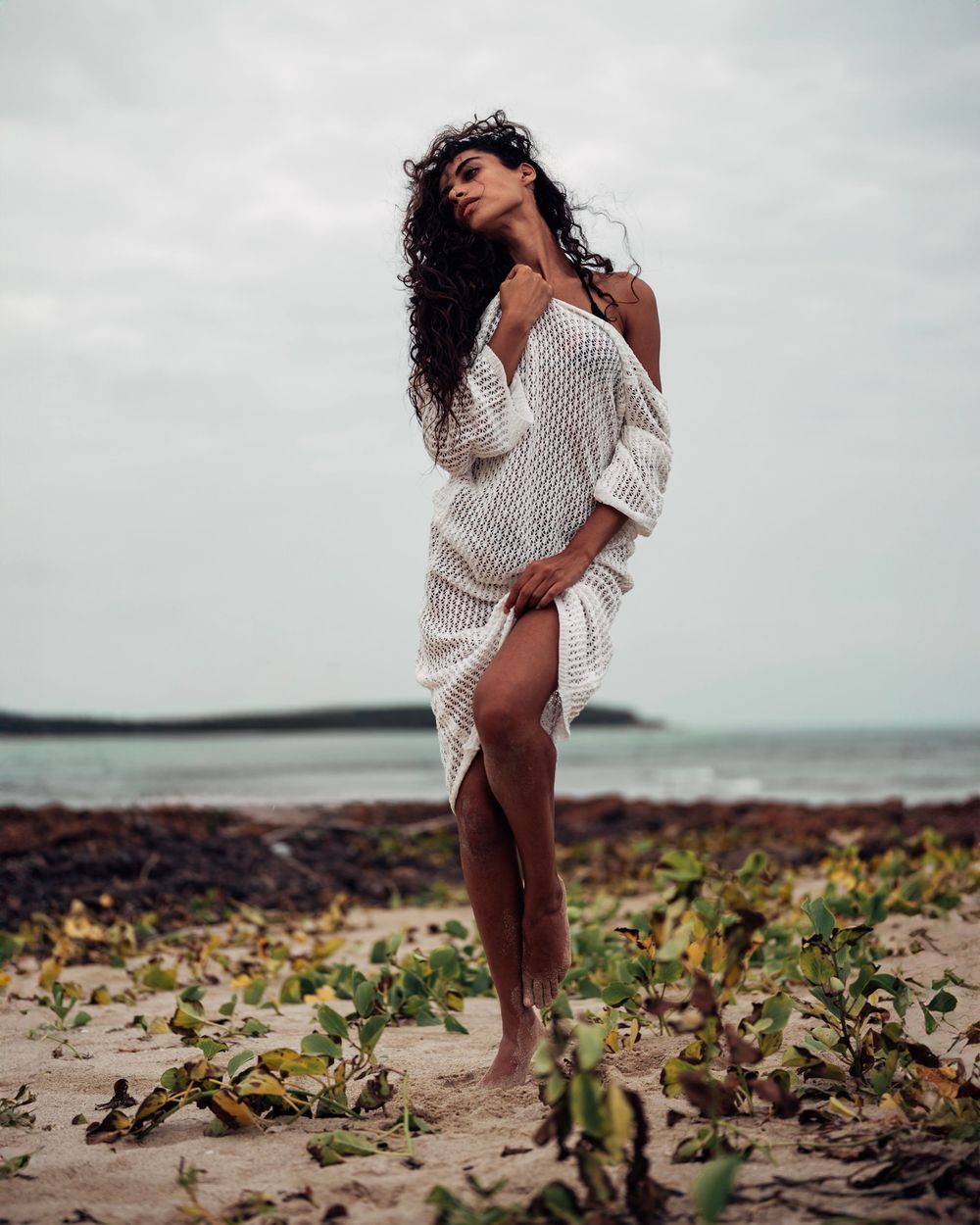 A series of photos showing someone in a white sweater dress posing on a cloudy beach with tropical foliage.