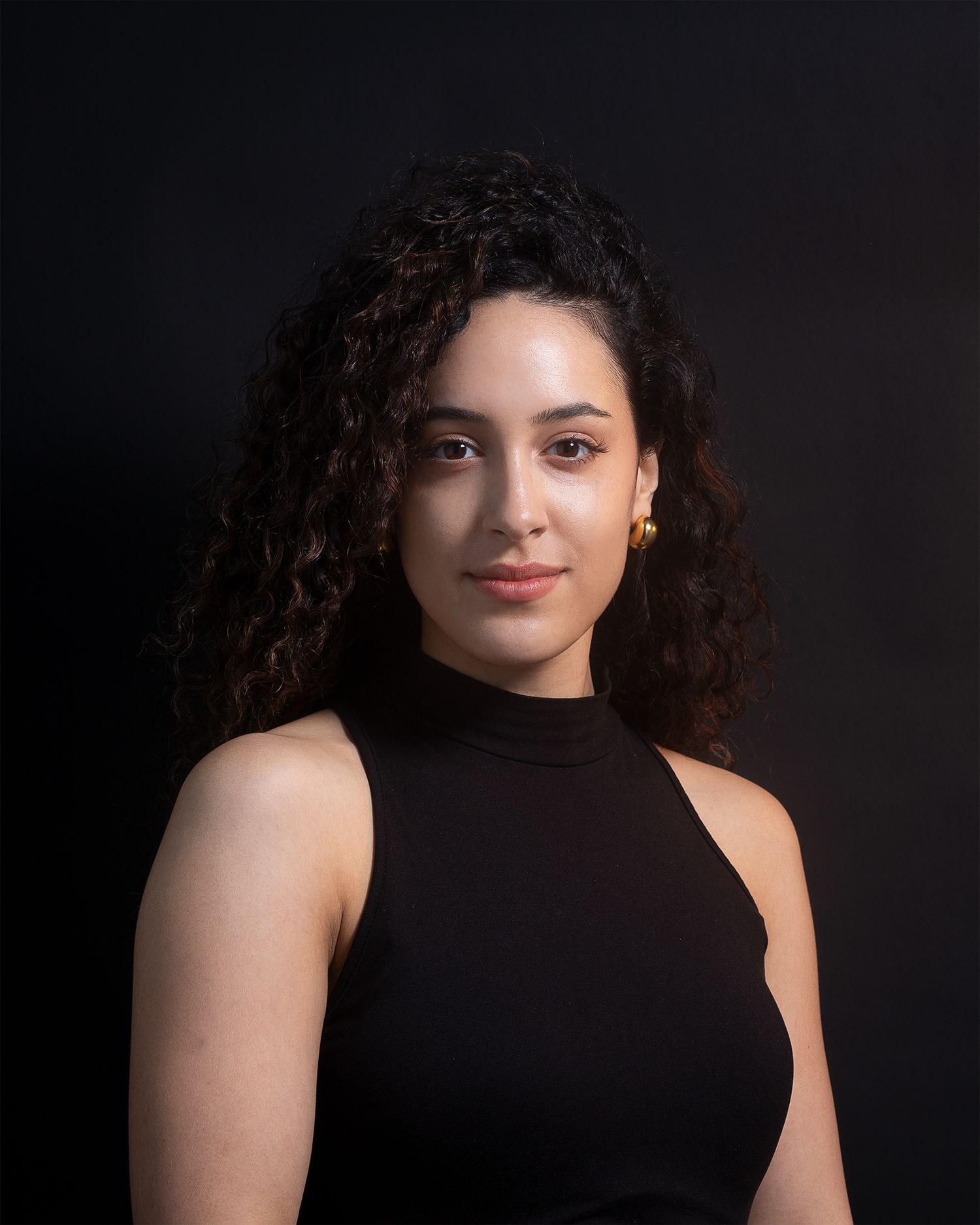 A studio portrait of a person with curly dark hair wearing a black sleeveless turtleneck.