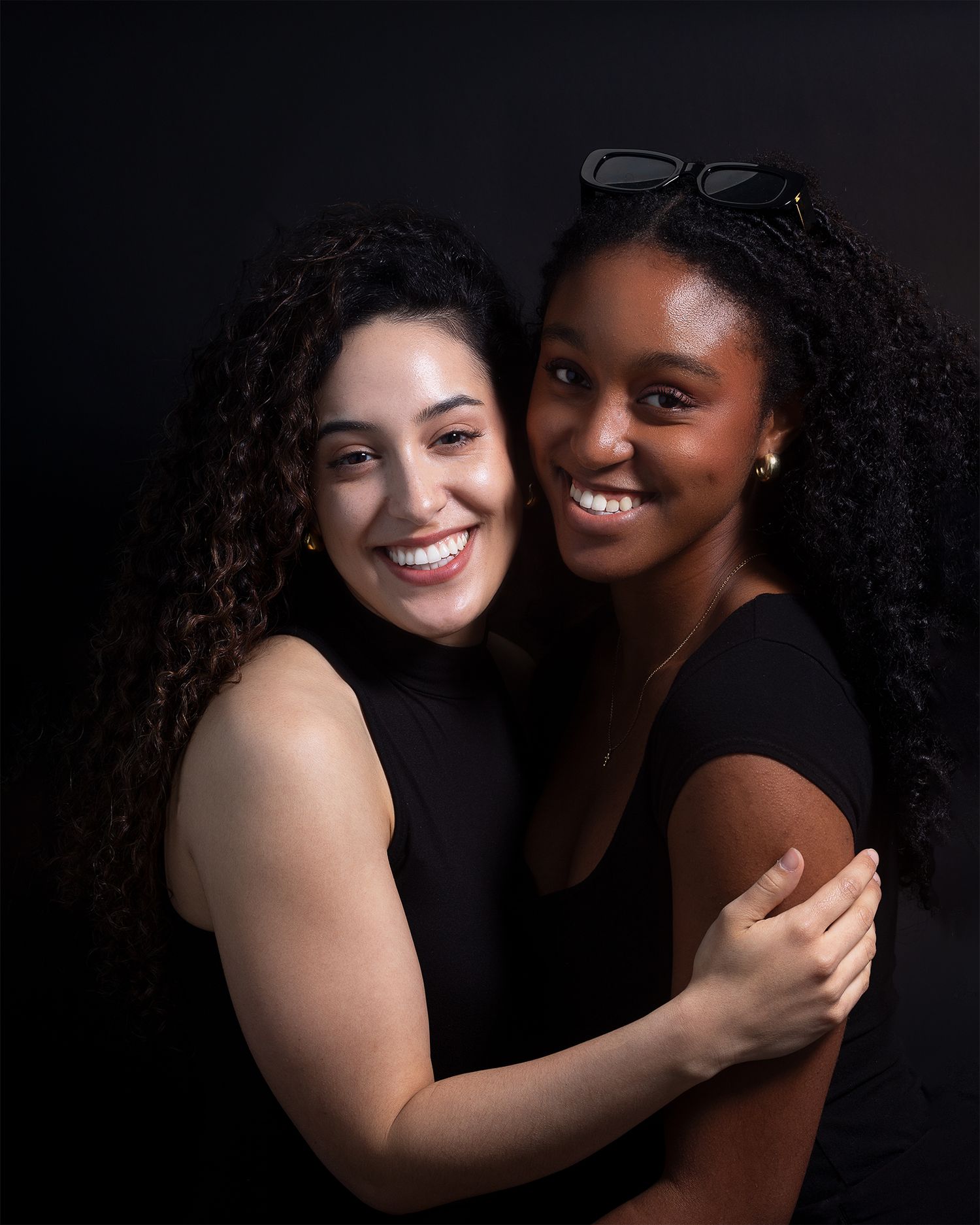 Two friends embrace while wearing black tops against dark background in studio portrait.