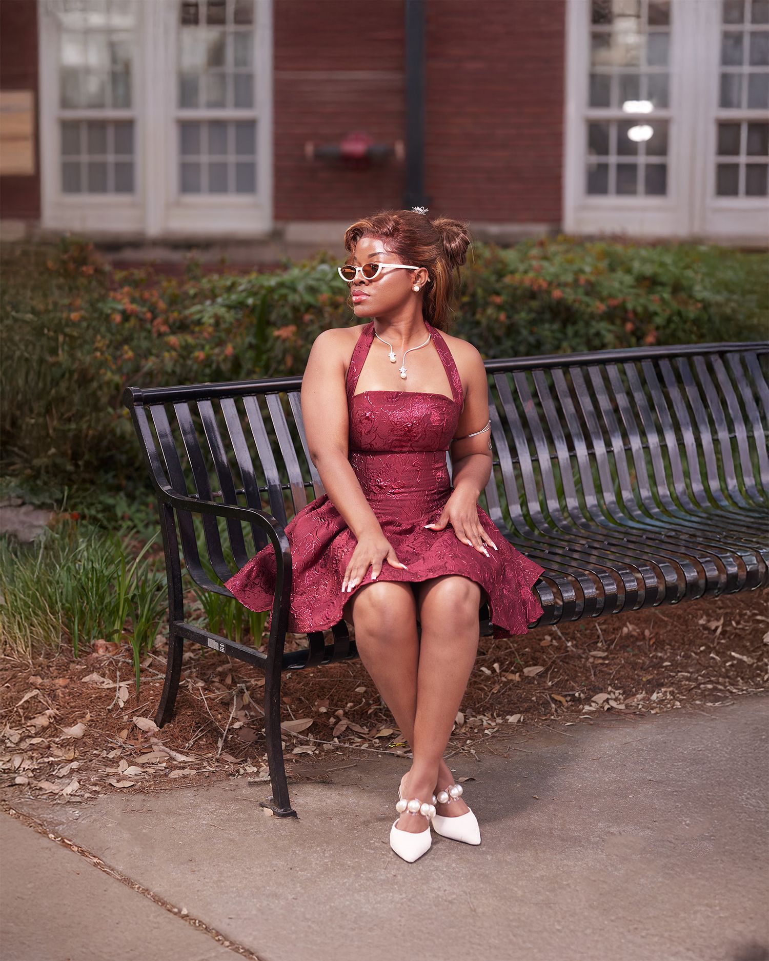 Woman in burgundy halter dress and white sunglasses sitting gracefully on a black park bench outdoors.