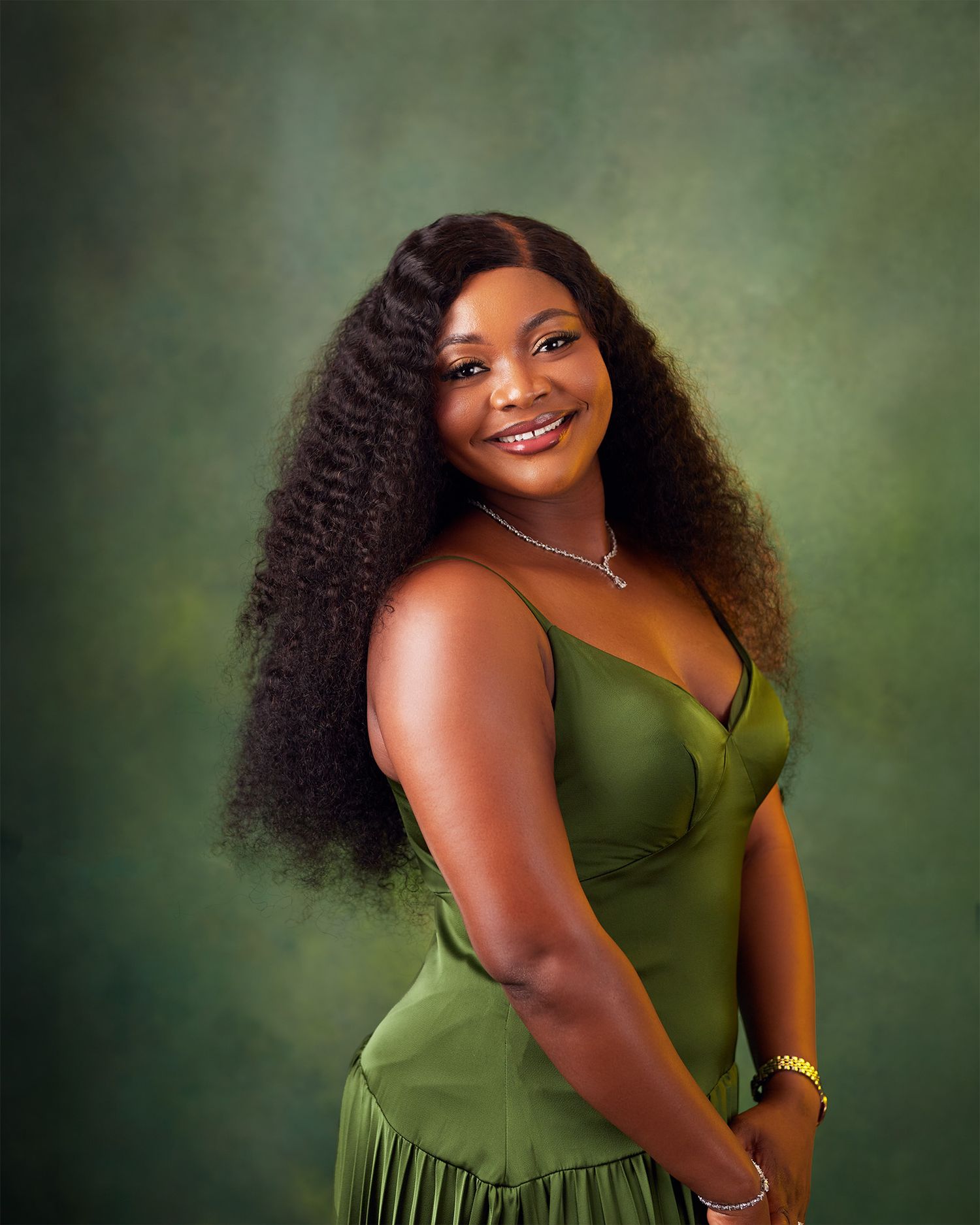 Woman in green satin dress with long curly hair smiling against a green studio background.
