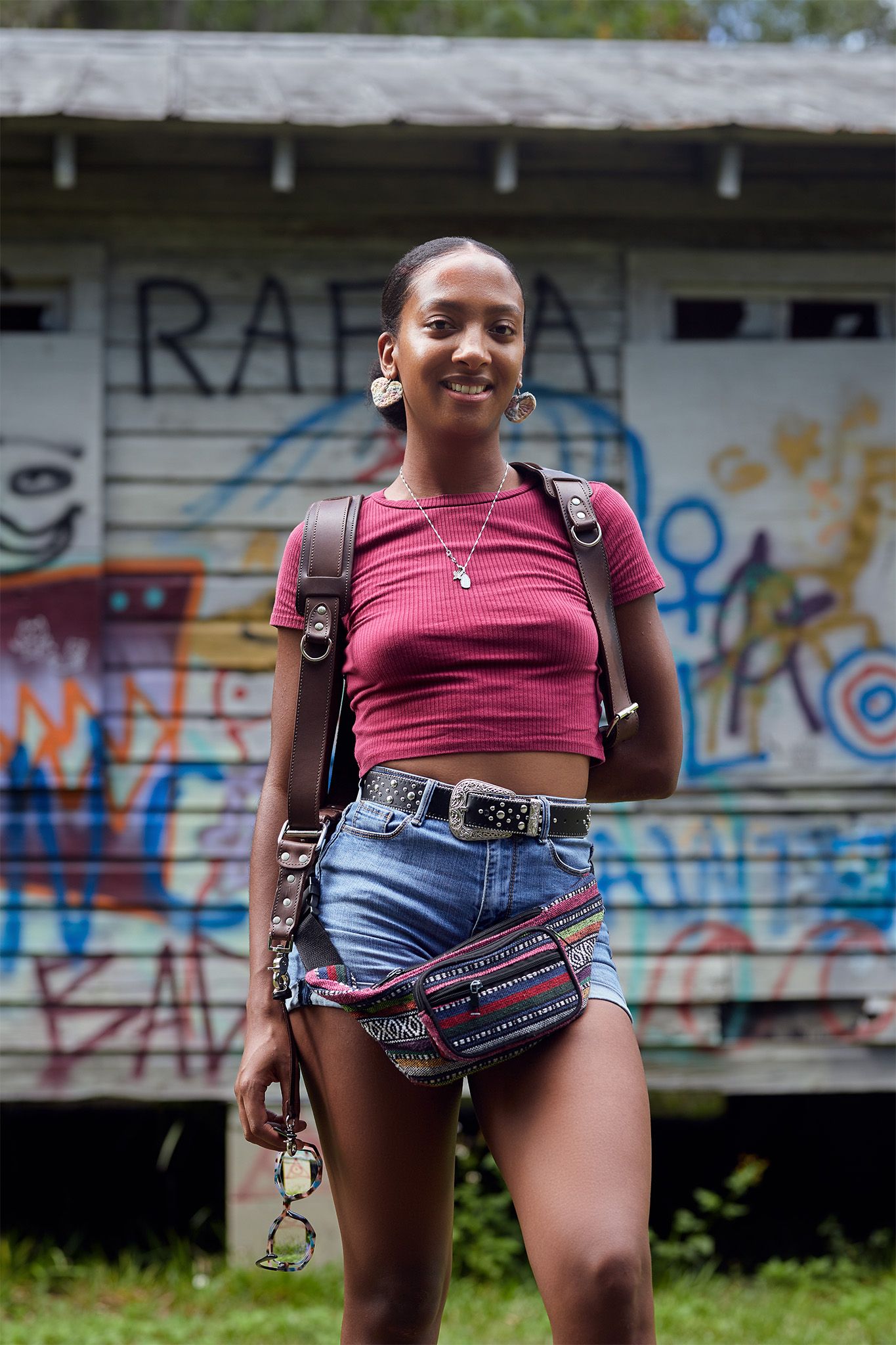 Young woman in red crop top and denim shorts smiling in front of graffiti-covered wooden building.