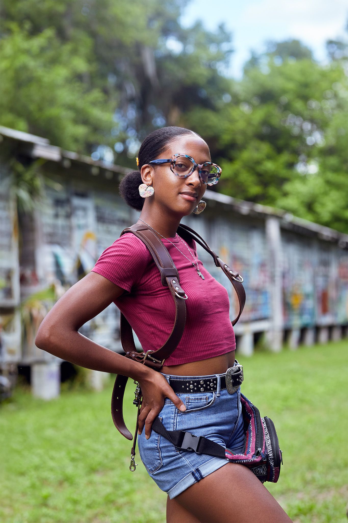 Young Black woman in red crop top, denim shorts, glasses, and leather backpack posing outdoors near graffiti wall.