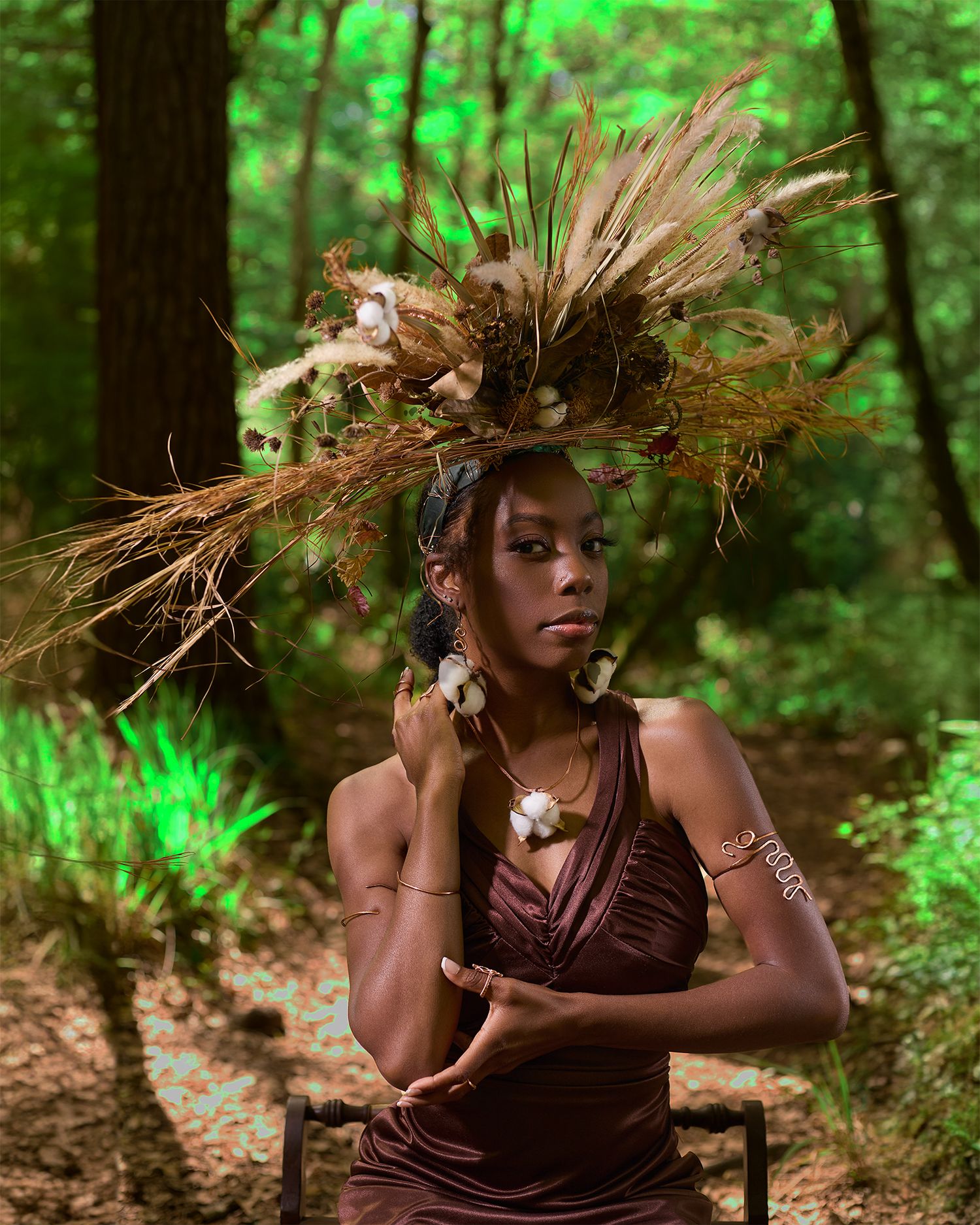 A nature-inspired fashion photo featuring an elaborate dried grass headpiece and brown dress in a forest setting.