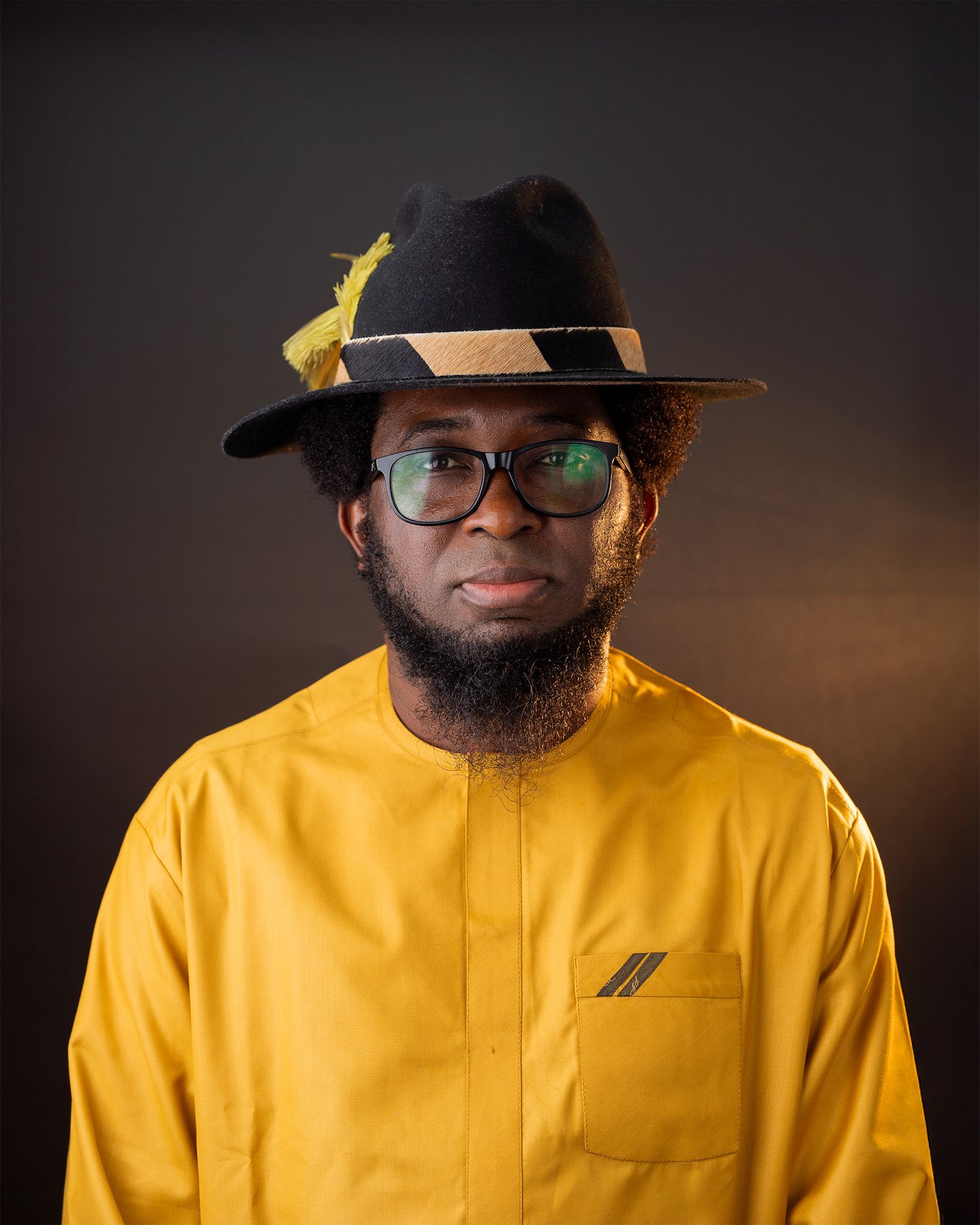 A person in a yellow top and black hat with glasses poses for a professional headshot against a dark backdrop.
