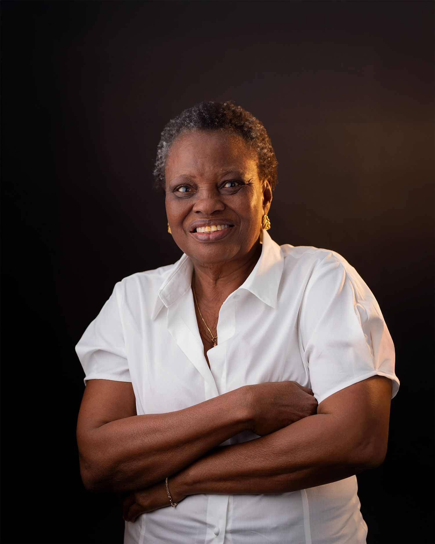 A smiling woman in a white button-down shirt poses with crossed arms against a dark backdrop for a professional portrait.