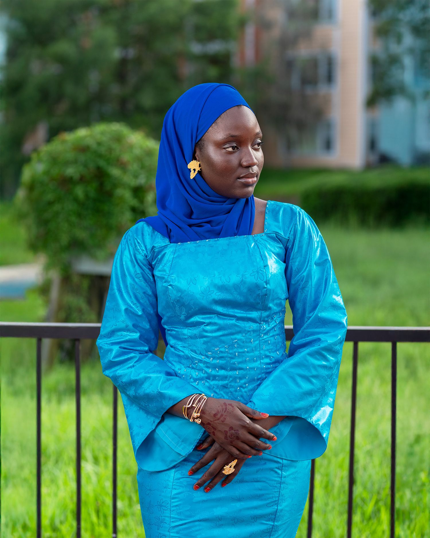 An elegant woman wearing a bright turquoise dress and royal blue hijab poses thoughtfully against a garden railing.