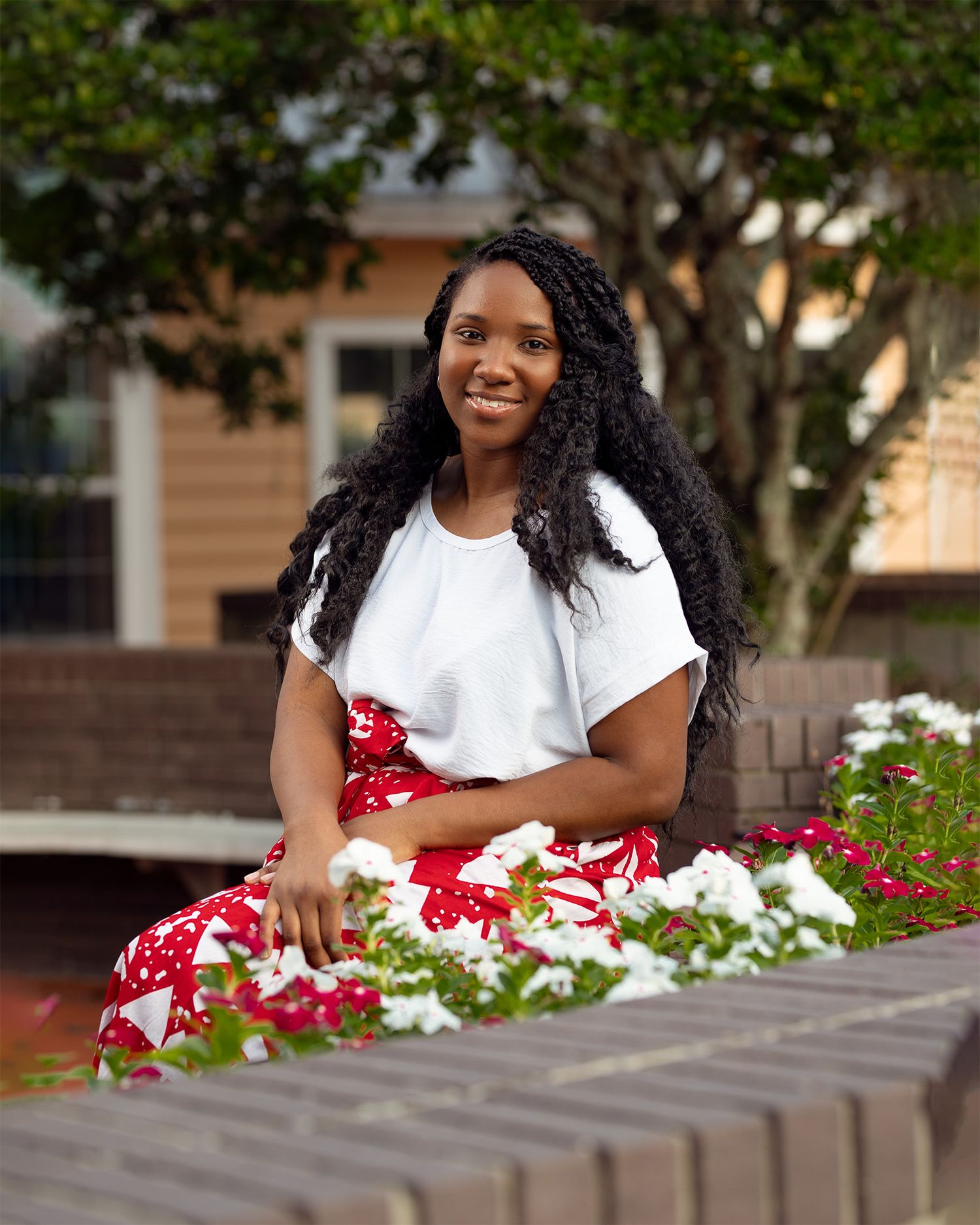 A person in a white t-shirt and red floral dress smiles while sitting outdoors on concrete steps near white flowers.