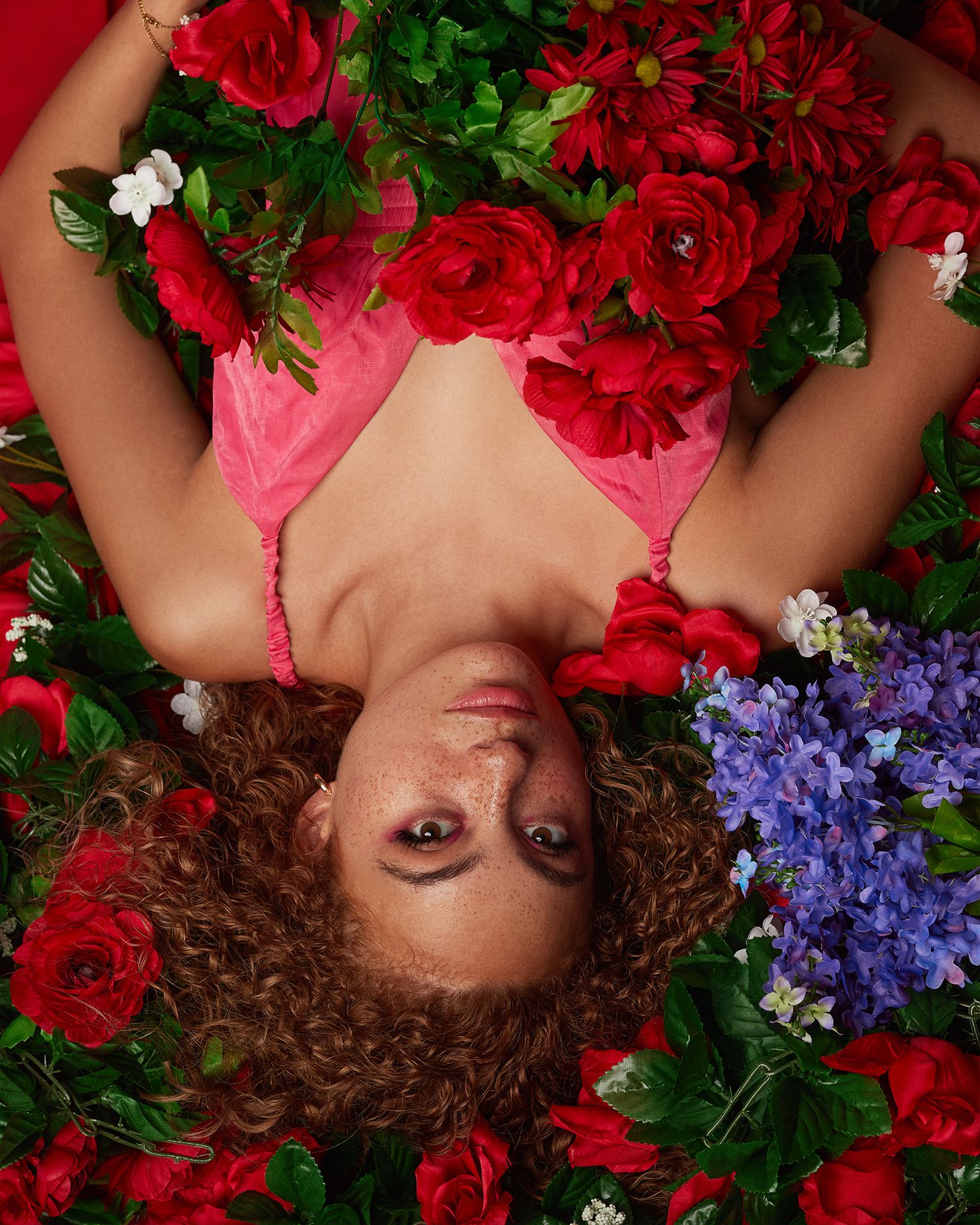 Artistic portrait surrounded by red roses and purple flowers against pink background.