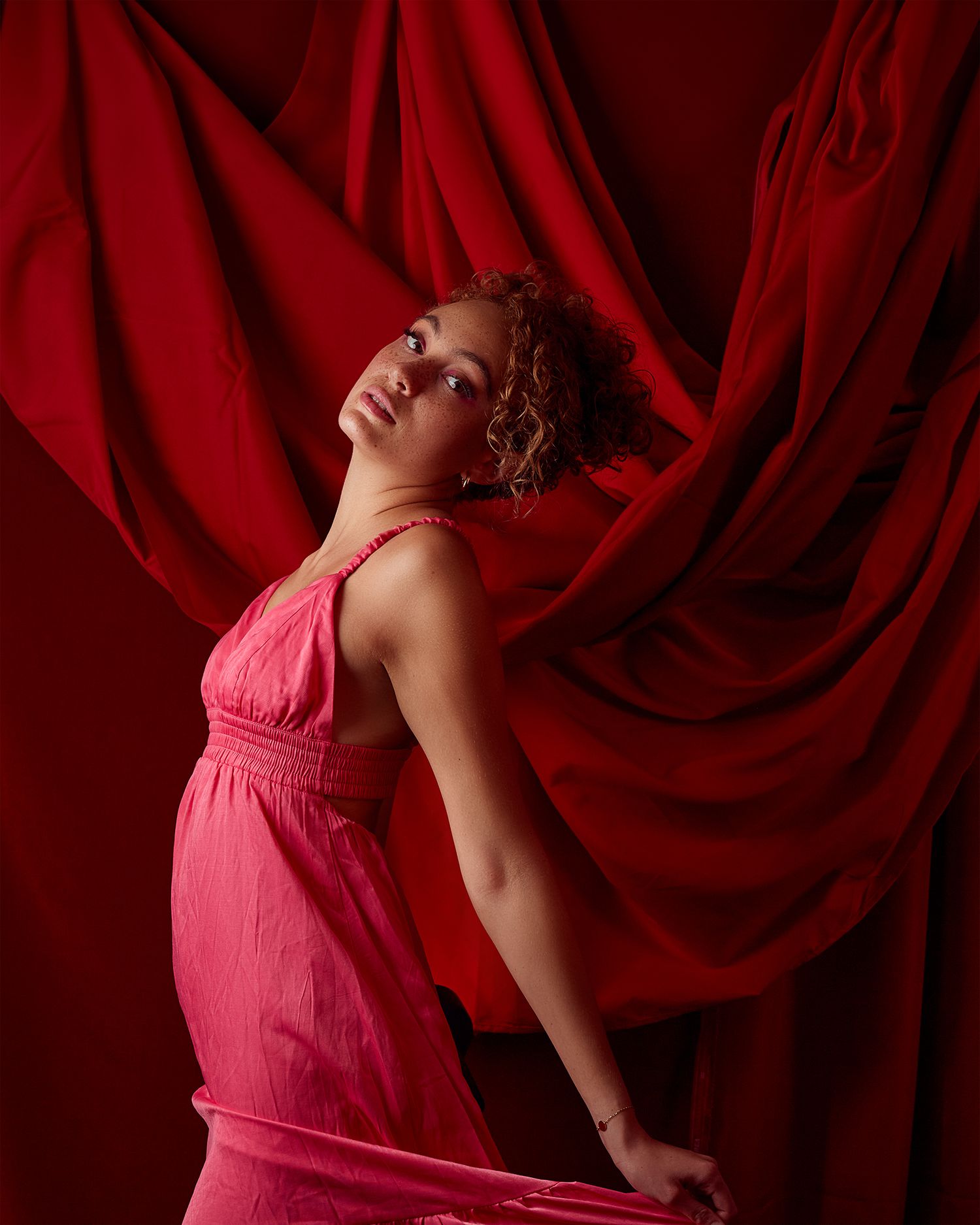 Dramatic fashion pose against red draped backdrop wearing pink dress.