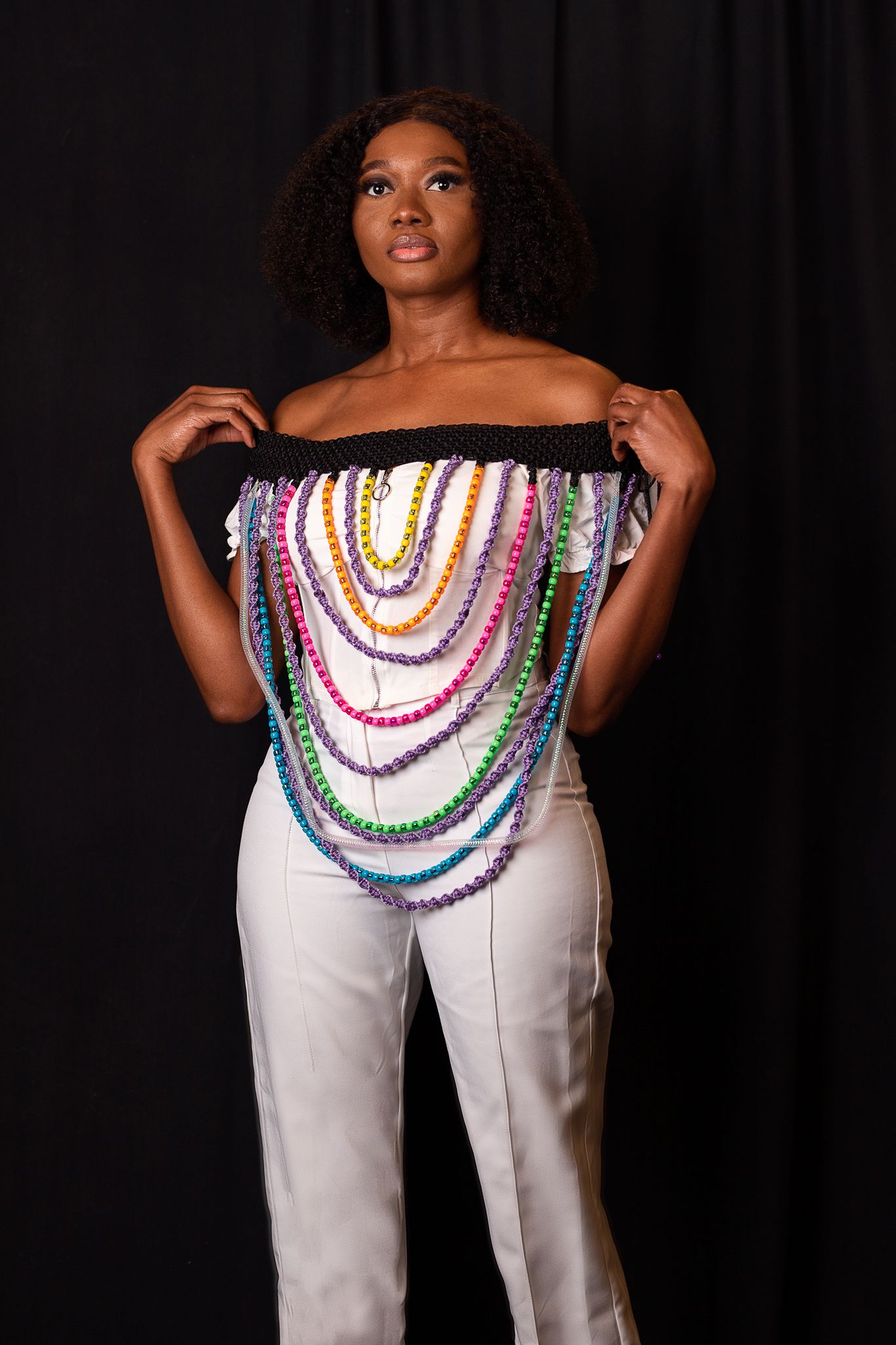 Fashion model in white pants and colorful beaded necklace design against dark background.