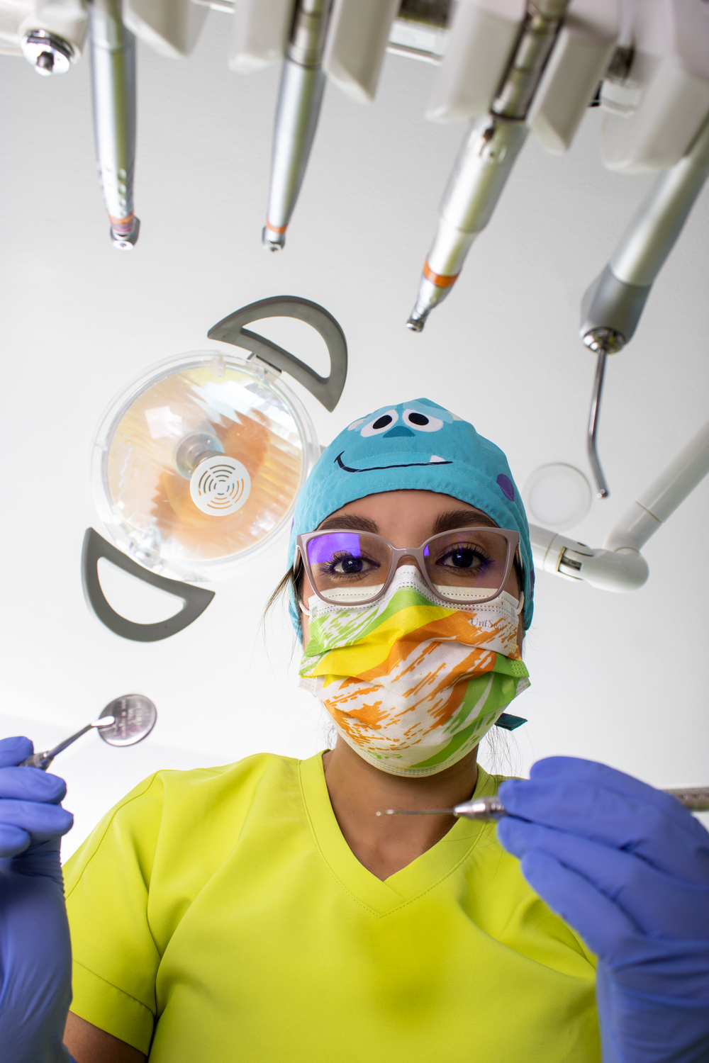 Dentist in colorful scrubs and mask holding dental tools viewed from patient perspective.