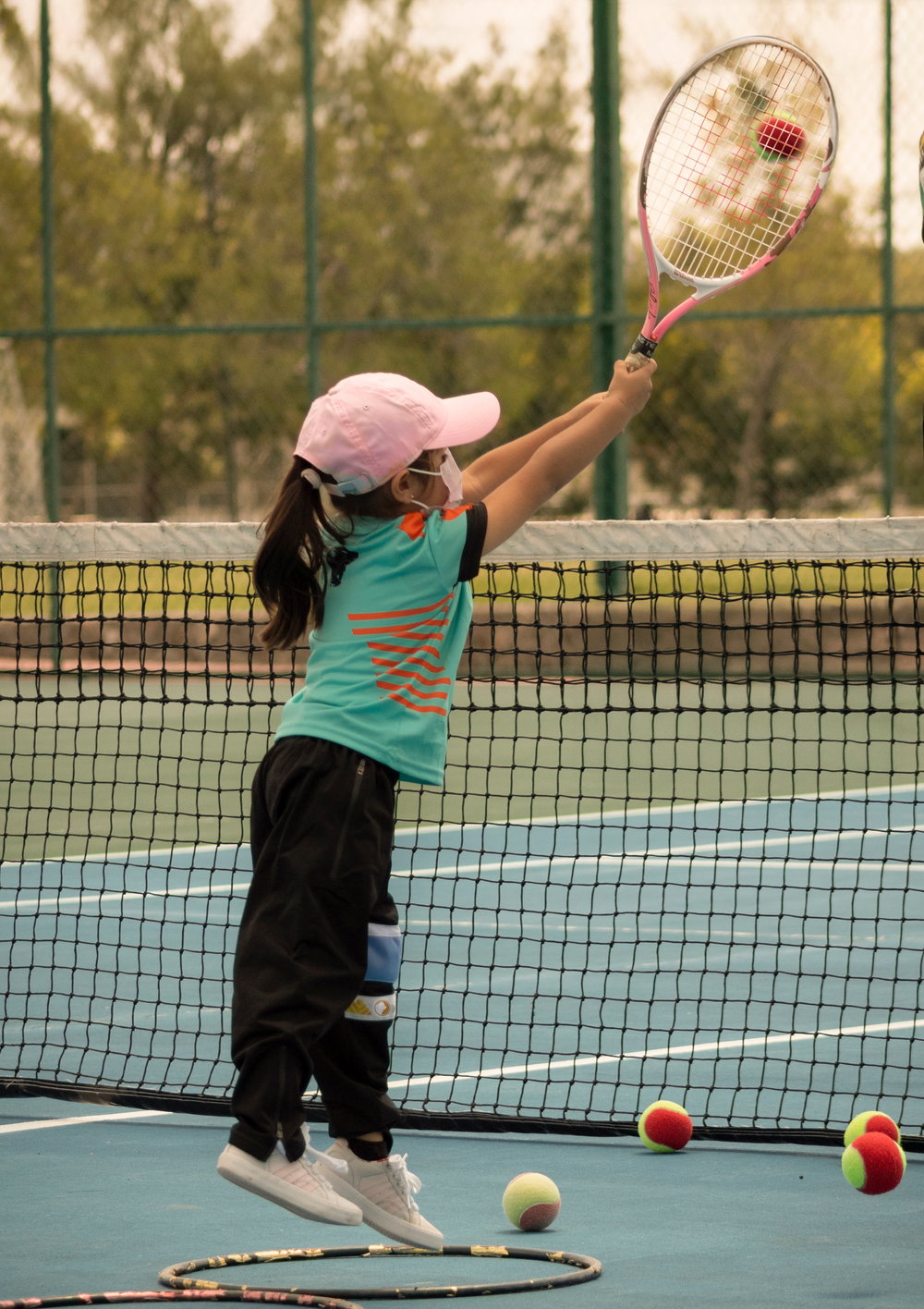 Young girl in pink cap and teal shirt practicing tennis swing at net on outdoor court.