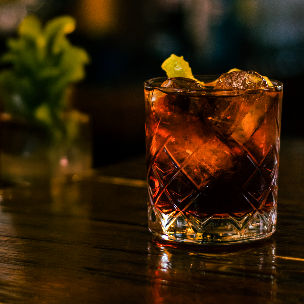 Dark whiskey cocktail with ice and citrus garnish in crystal glass on wooden bar.