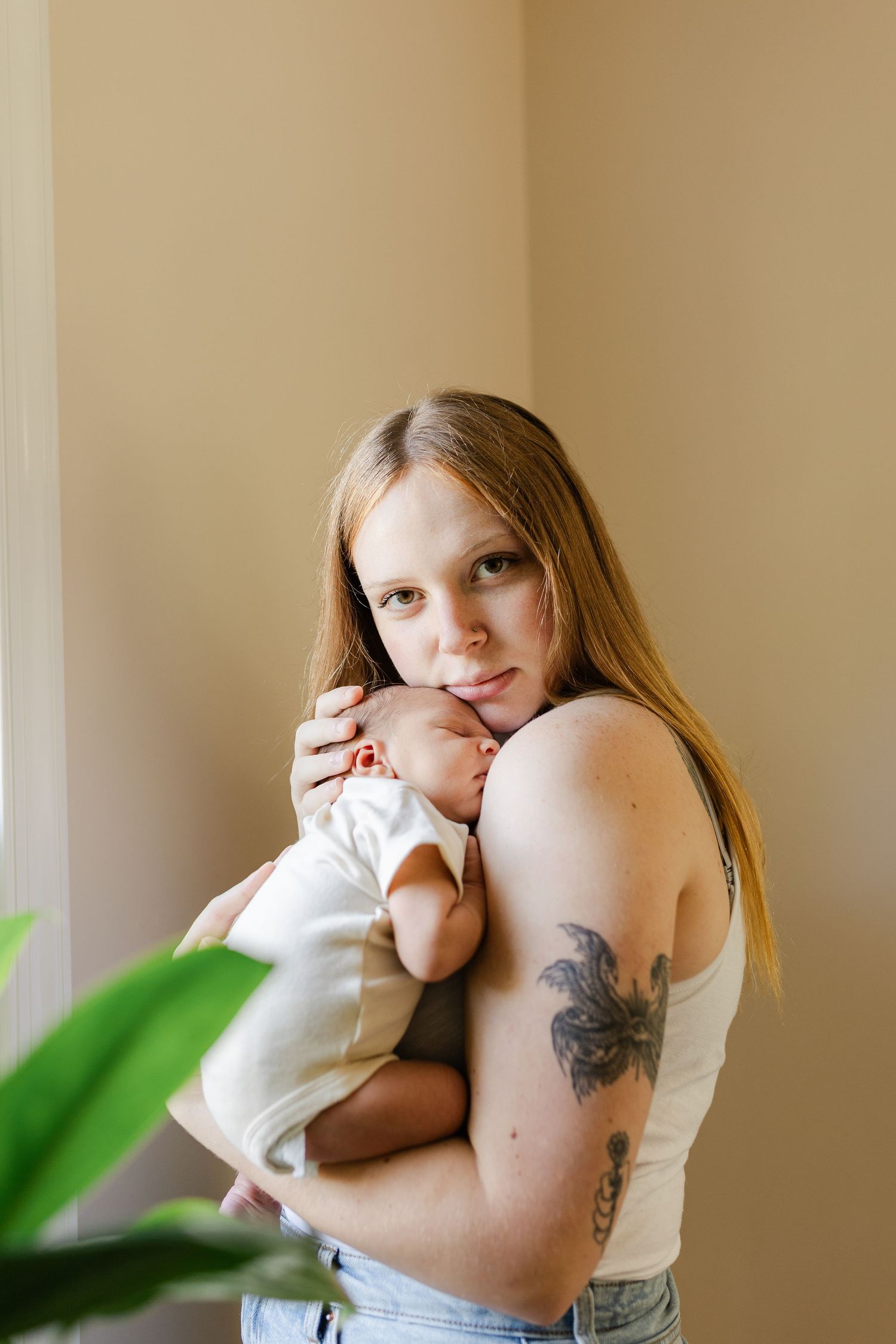 A couple shares a tender moment with their newborn baby in a sunlit room with houseplants.