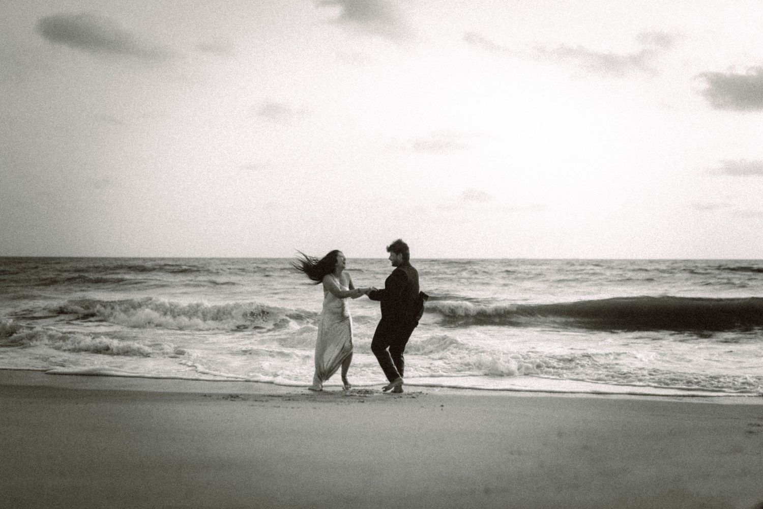 Black and white beach photoshoot with silhouettes walking along the shoreline at dusk.