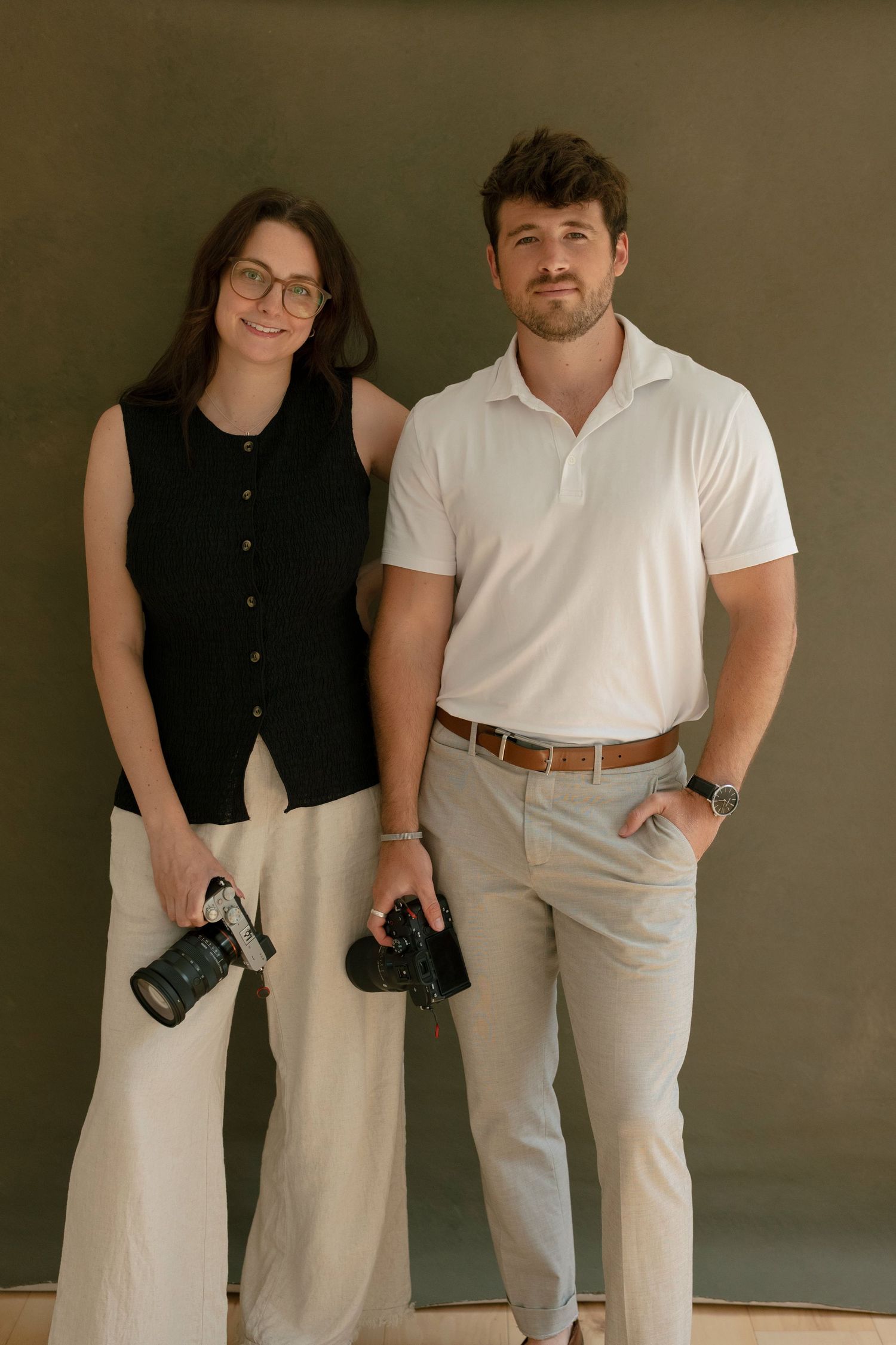 Male and female wedding photographers posing with cameras against olive green studio backdrop.