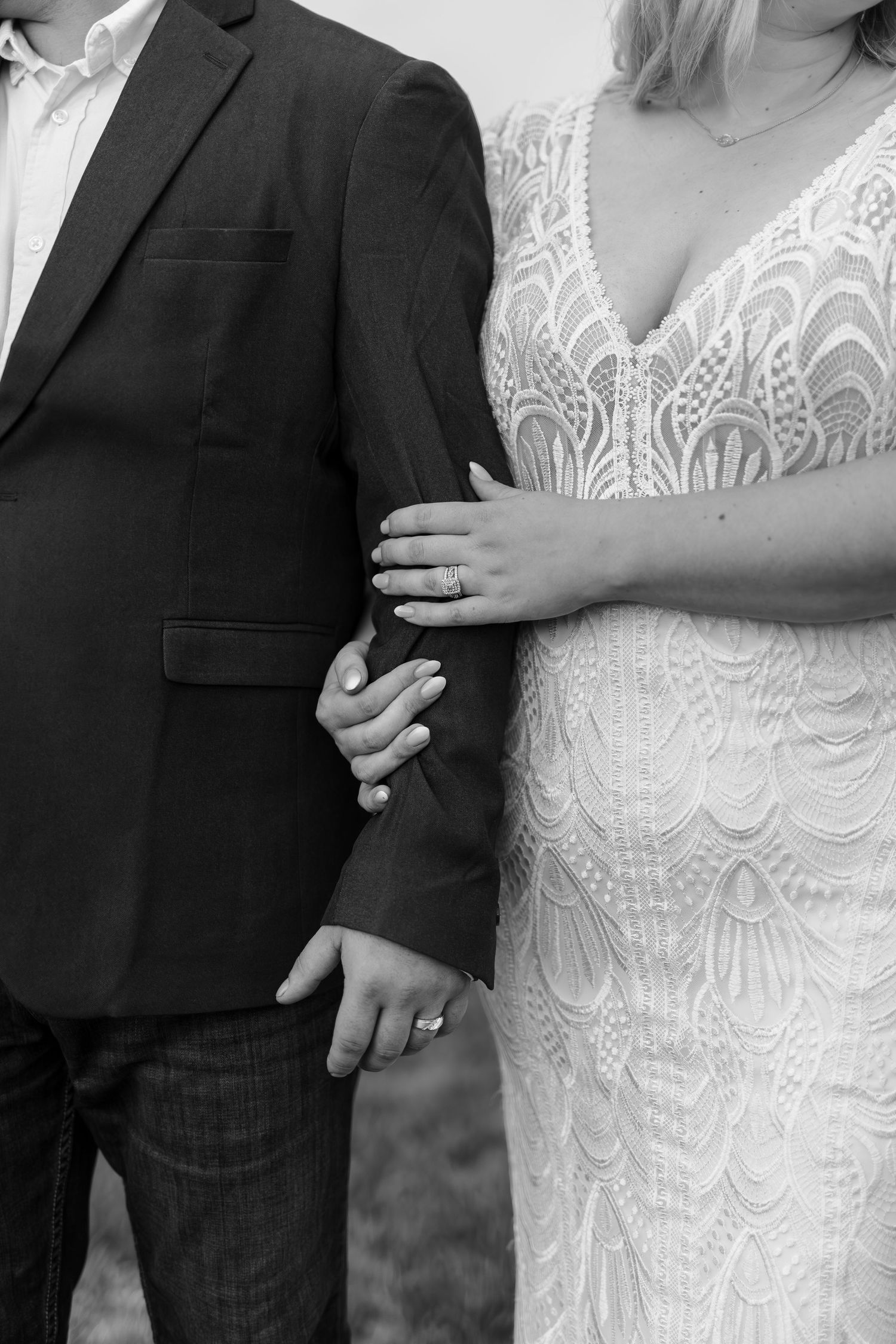 Black and white close-up of bride and groom holding hands, showcasing wedding rings and lace gown.