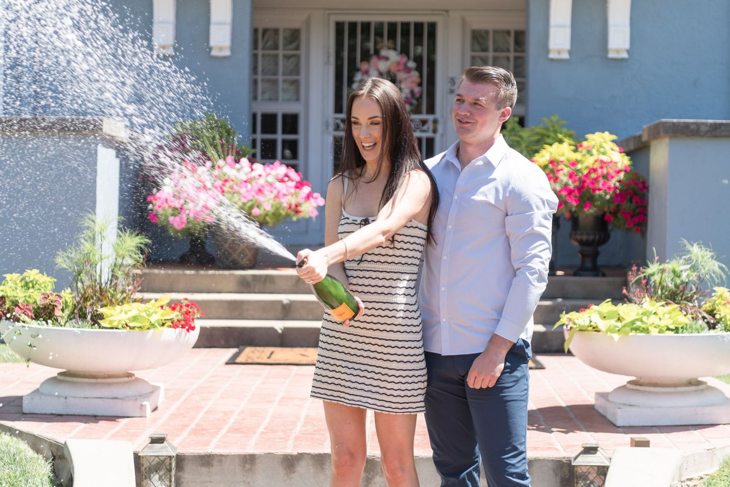 Couple pops champagne in front of blue house with colorful flower garden.
