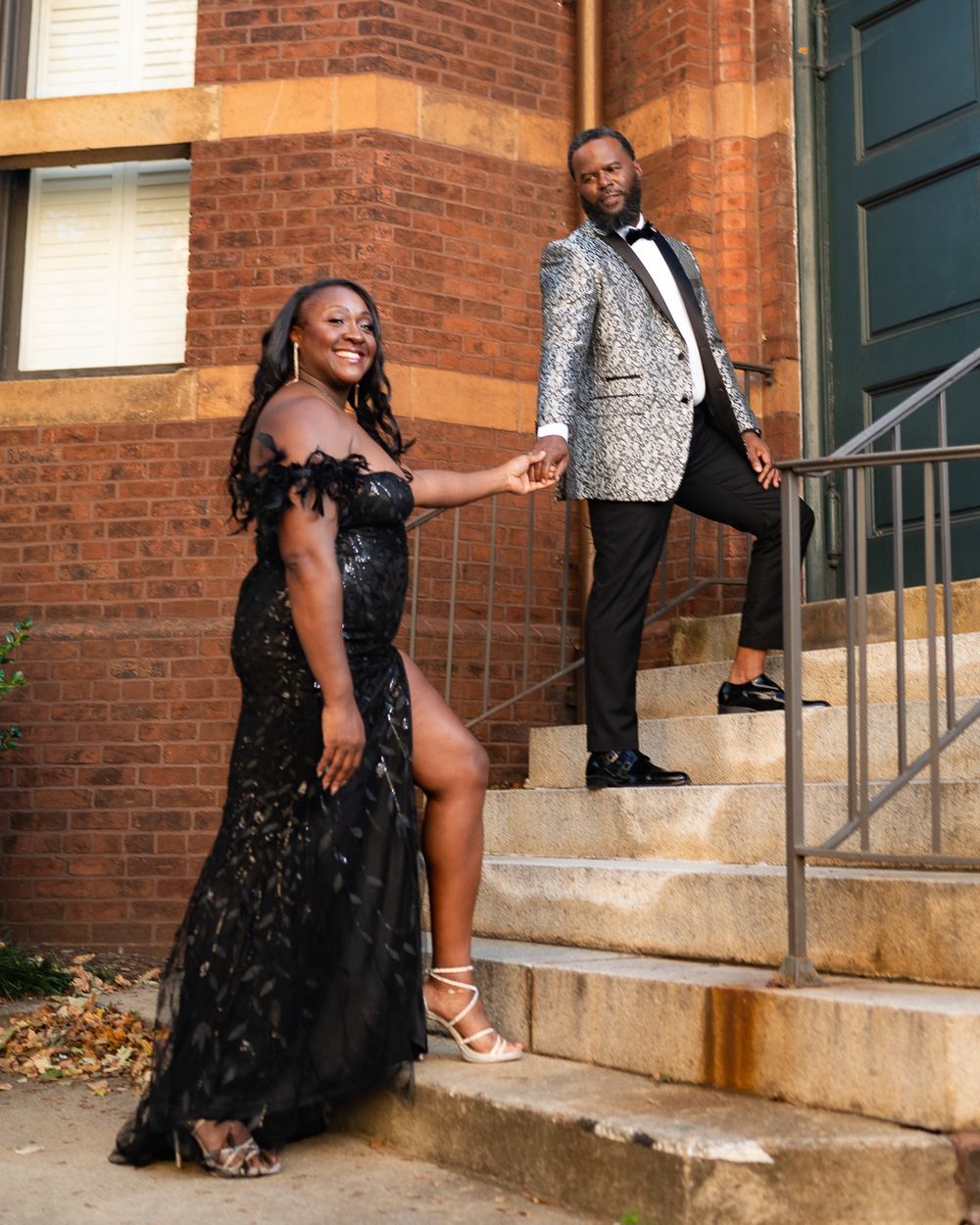 Formal couple poses on brick building steps, wearing black gown and silver tuxedo jacket.