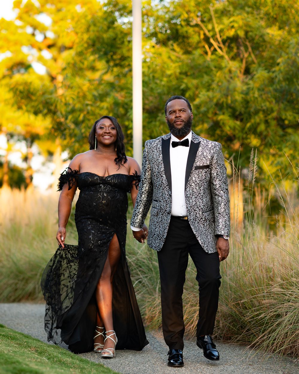 Couple in formal evening wear walking on path with autumn foliage backdrop.