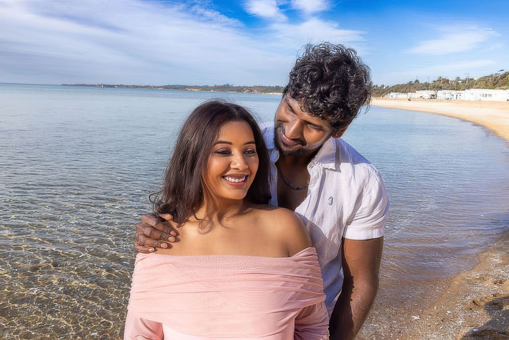 A couple shares an intimate moment together on a sunny beach with calm waters and sandy shoreline in the background.