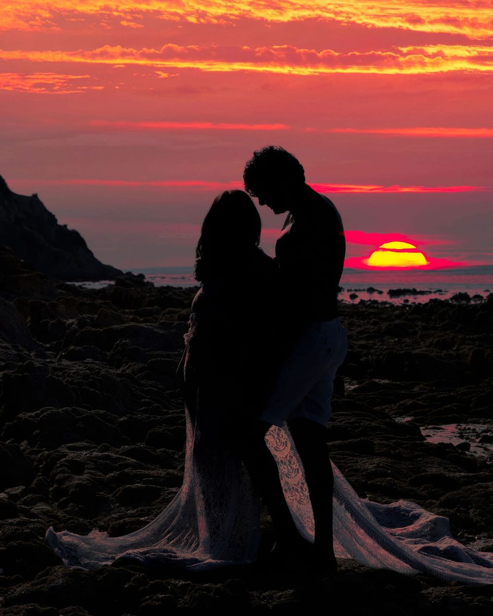 Silhouettes of figures against a vibrant orange and red sunset over a rocky coastal landscape at dusk.