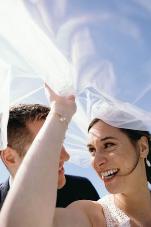 A joyful bride and groom share a tender moment under flowing white wedding veil against a blue sky background.
