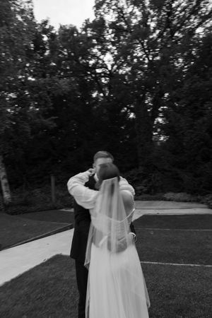 A black and white photo shows a formal couple in a romantic embrace outdoors, with the bride wearing a flowing veil.