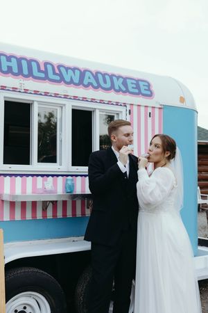 A couple in wedding attire poses together in front of a pink and blue striped ice cream truck called Chillwaukee.
