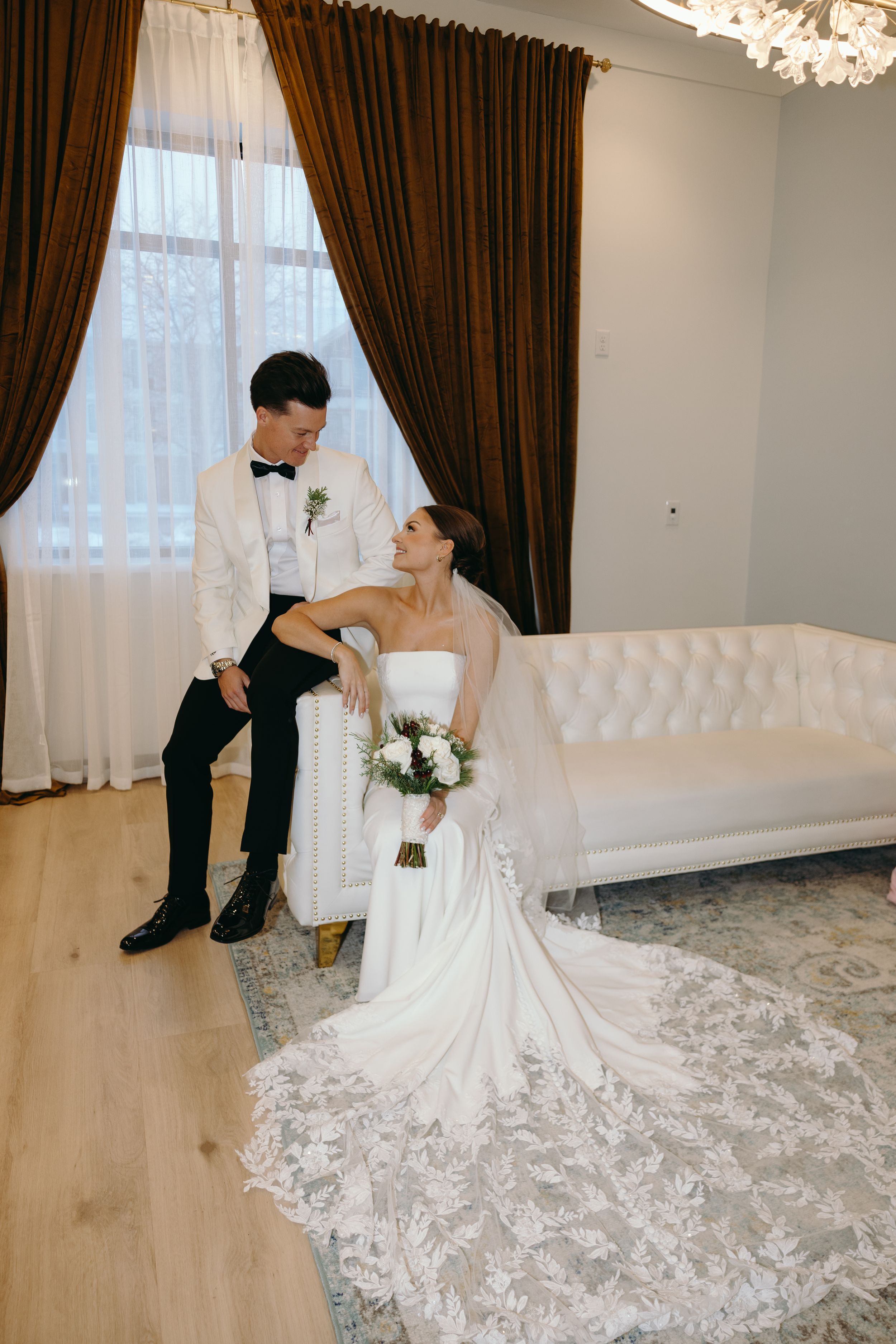 A bride in a stunning white lace wedding gown with a dramatic long train poses elegantly in a light-filled room.