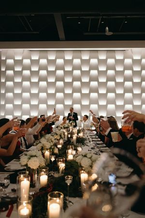 Wedding guests raise glasses in a toast at an elegant candlelit reception with white floral centerpieces and moody lighting.